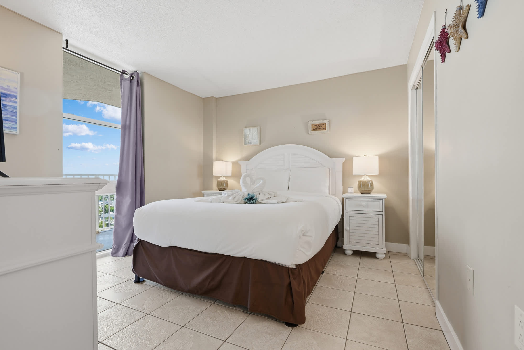 Dunes E1702 – Bright bedroom with a queen bed, lamps, and window view of blue sky.