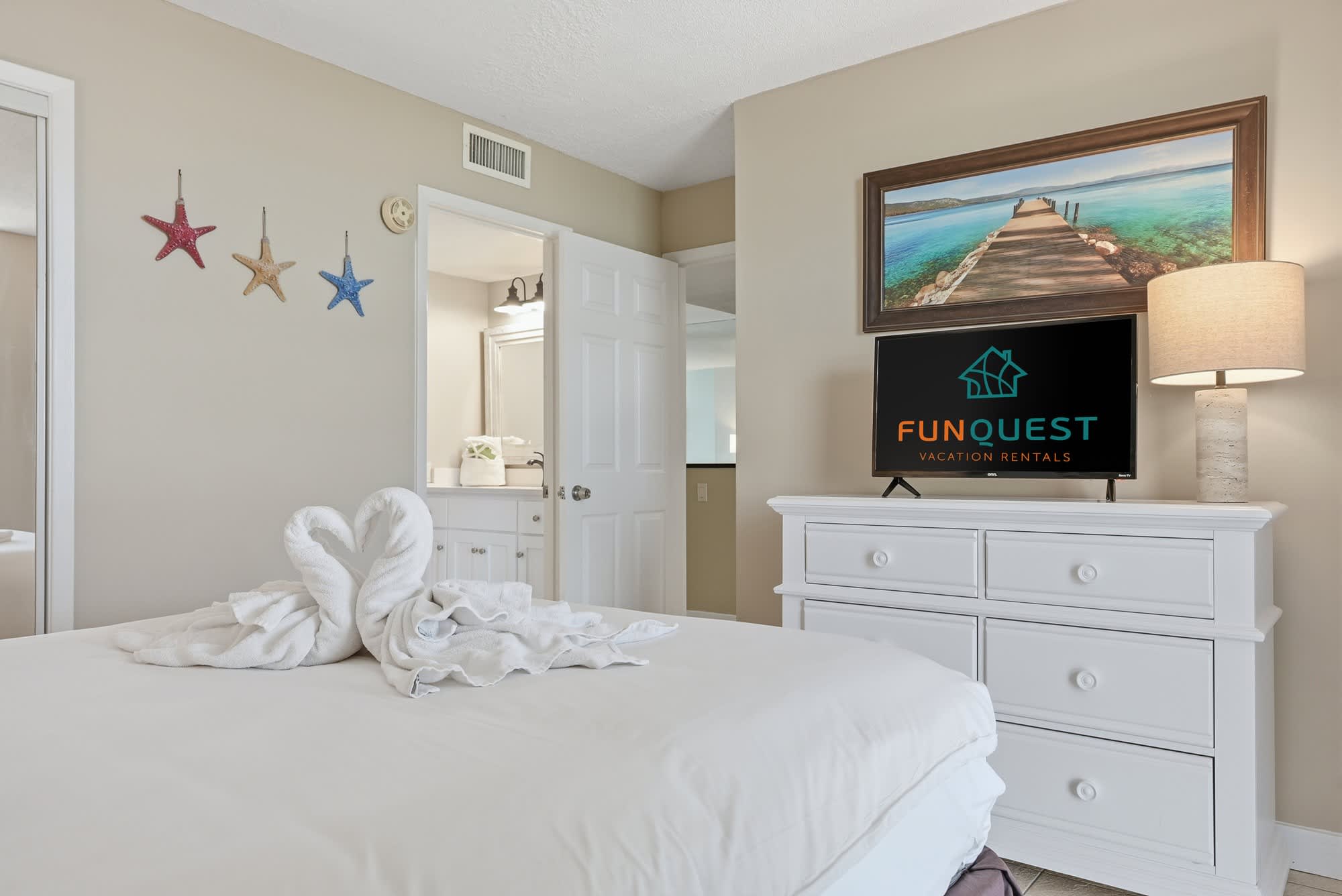 Dunes E1702 – Bedroom with a white bed, TV, dresser, and beach-themed decor including starfish.
