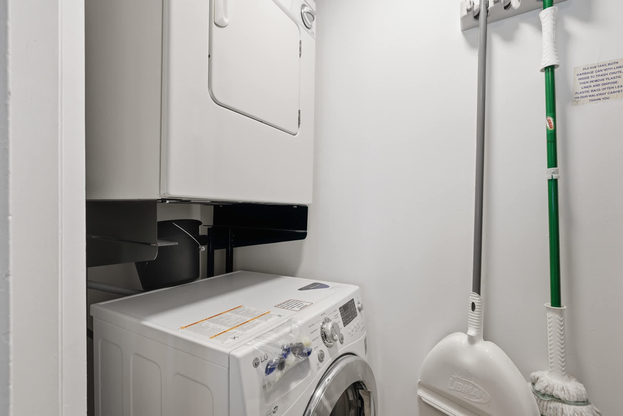Dunes E1702 – Laundry room with stacked washer and dryer, broom, dustpan, and white walls.