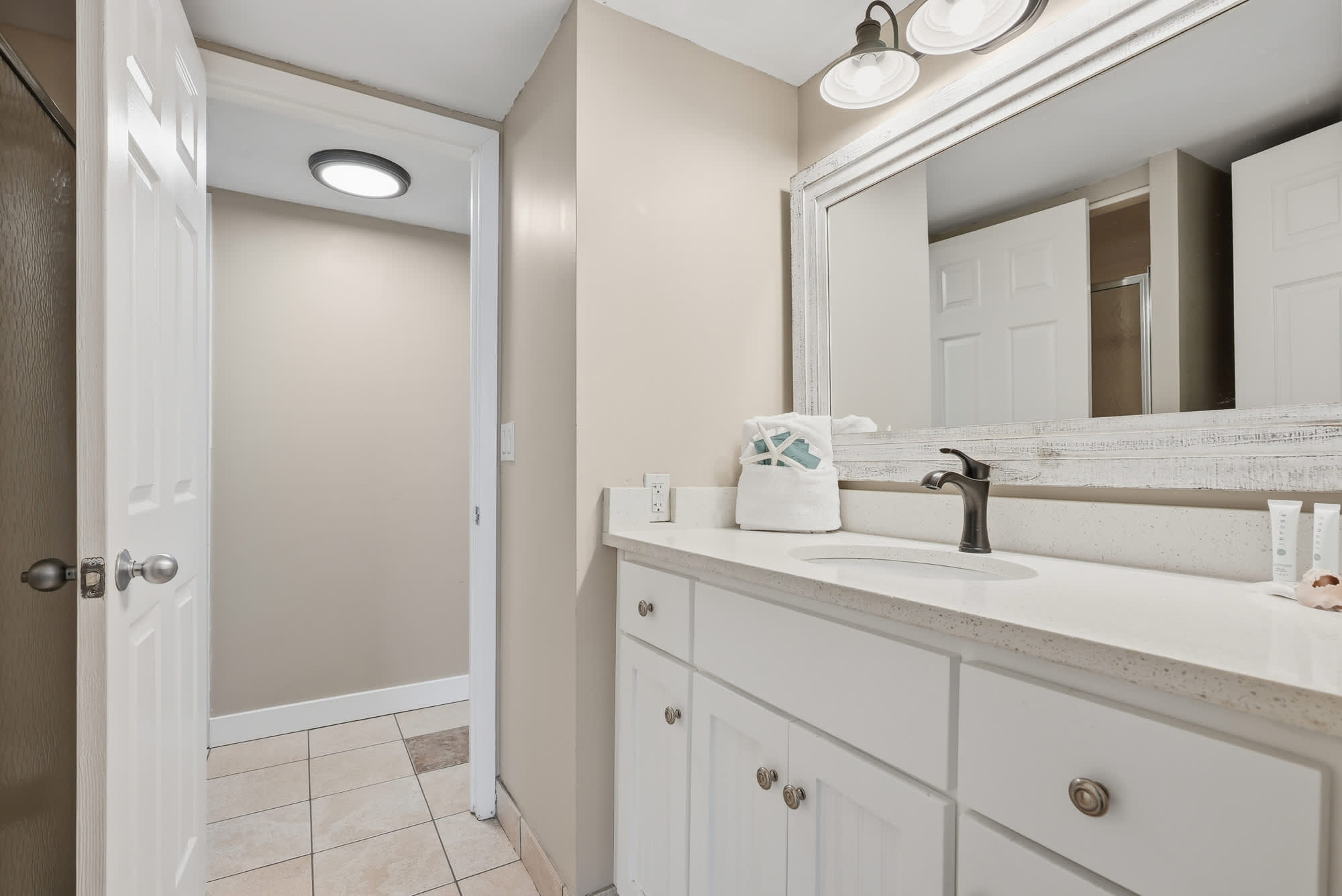 Dunes E1702 – Bathroom with double vanity, mirror, and light fixtures; tile flooring and open door leading to another room.