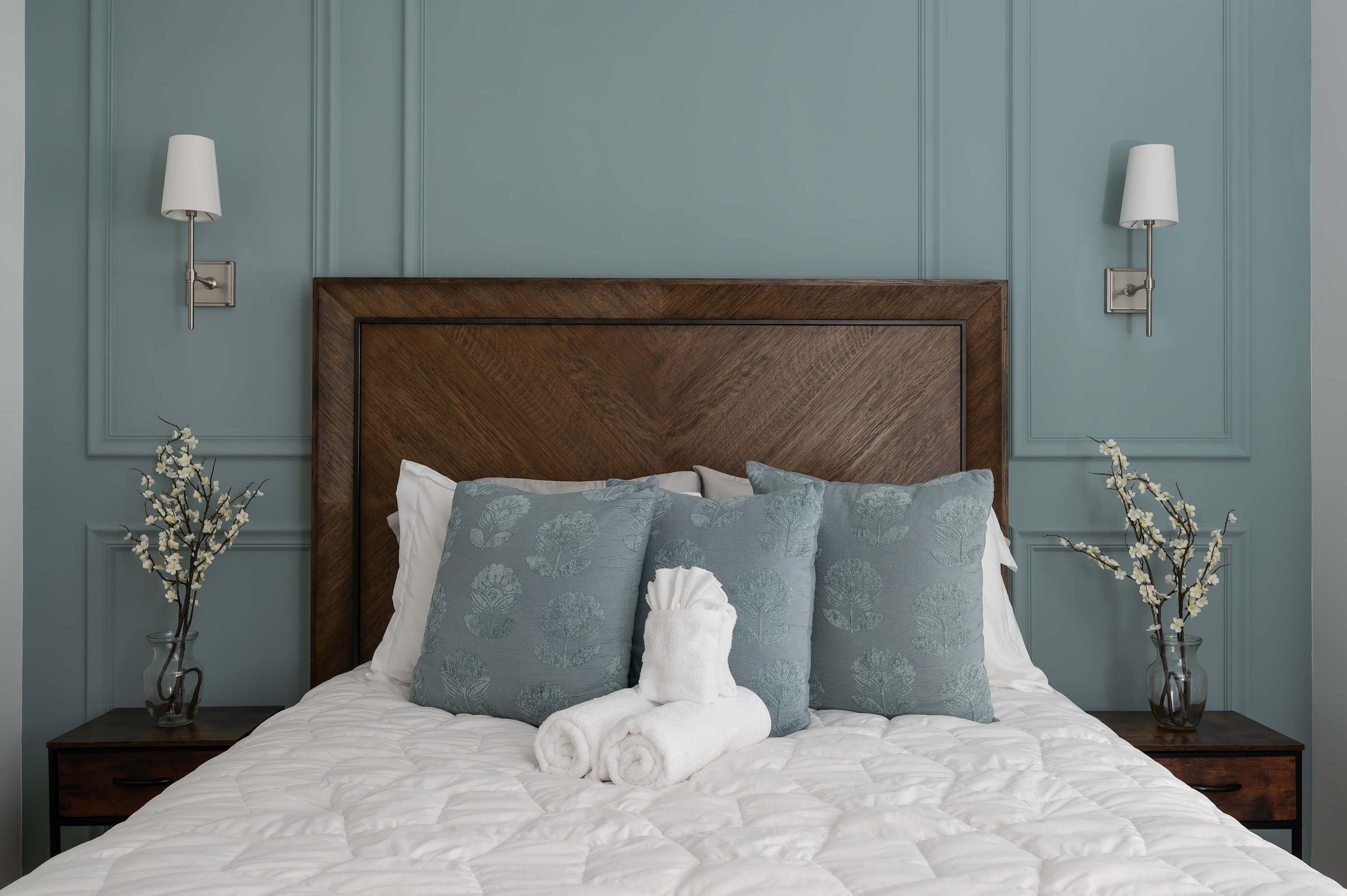 Cozy bedroom featuring a wooden headboard, soft blue pillows, and elegant wall sconces for a relaxing atmosphere.