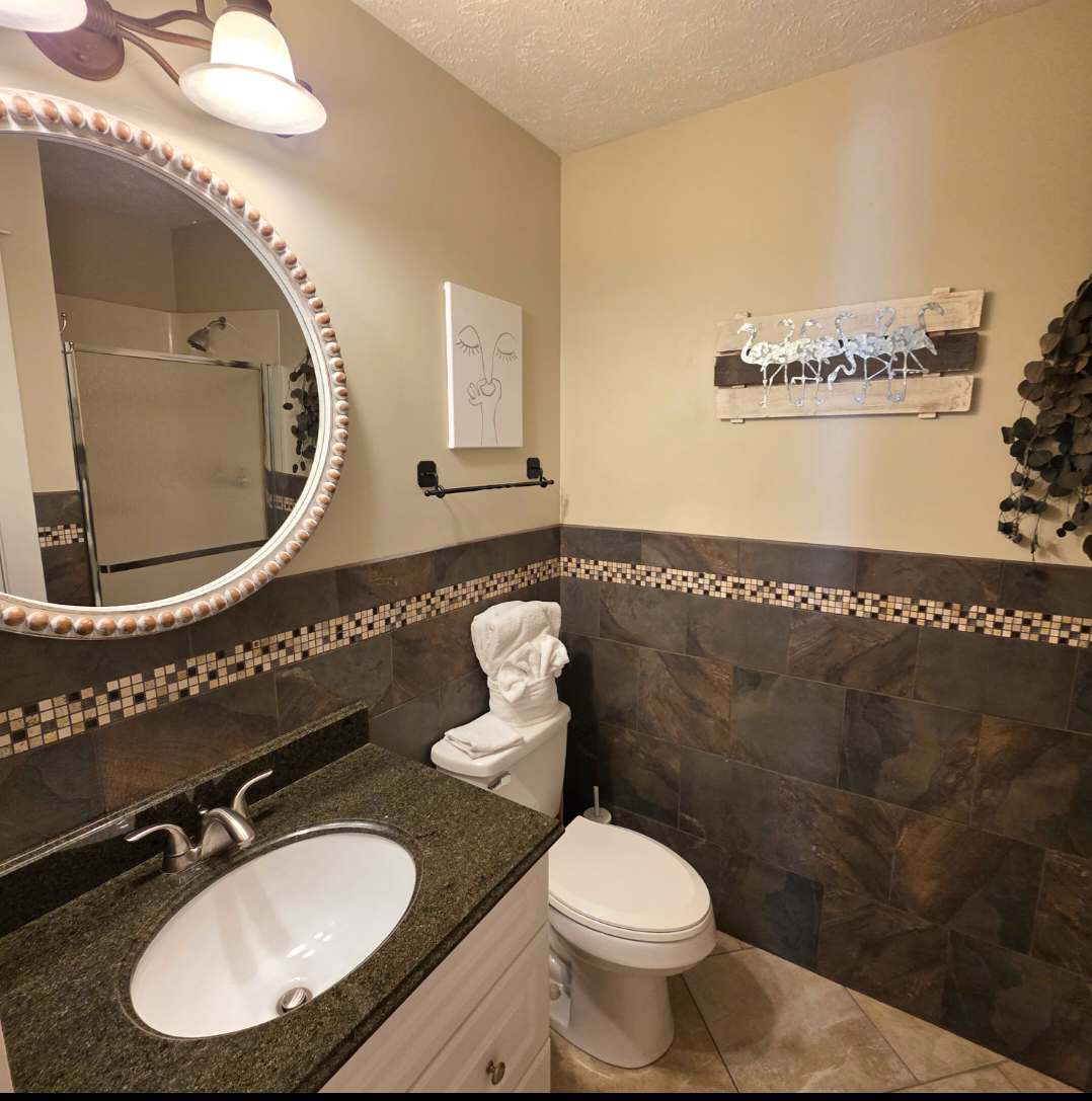 7405 Beach Dr A – Bathroom with dark stone tiles, a circular mirror, sink, toilet, and decorative wall art.