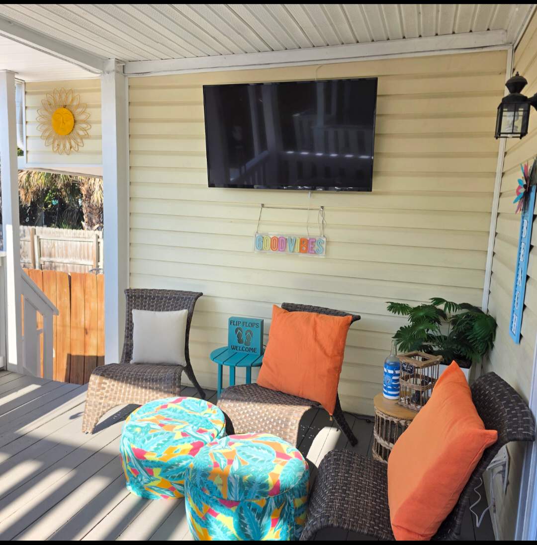 7405 Beach Dr A – Covered porch with seating, flat-screen TV, colorful poufs, and potted plants.