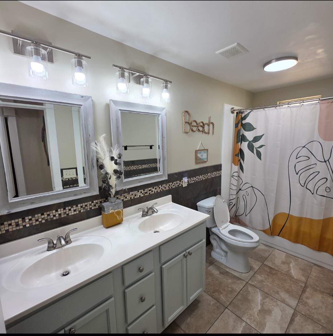 7405 Beach Dr A – Bright bathroom with double sinks, decorative mirrors, tile floor, and a shower curtain featuring leaf designs.