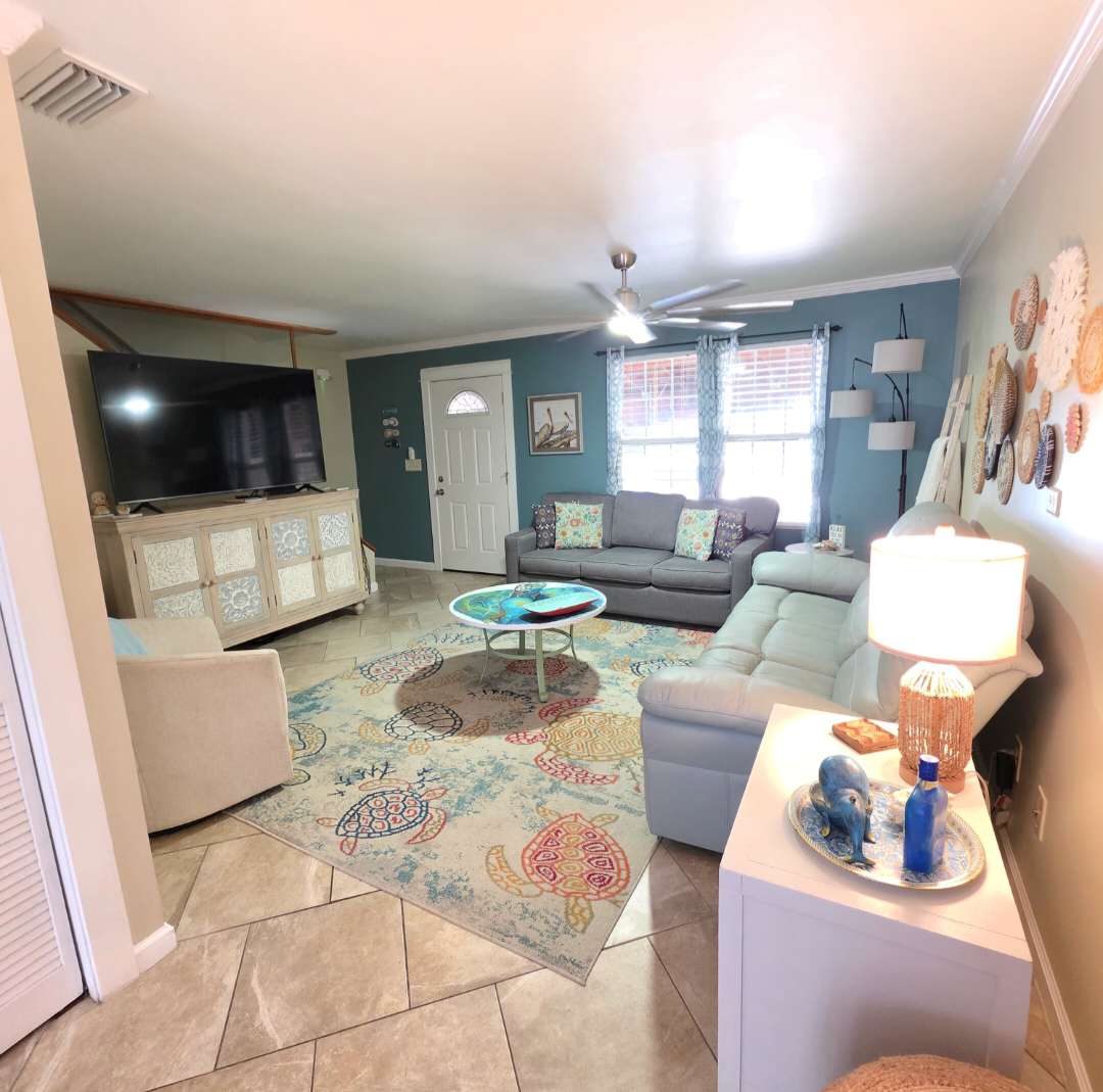 7405 Beach Dr A – Living room with gray sofas, large TV, decorative rug, and colorful wall art.