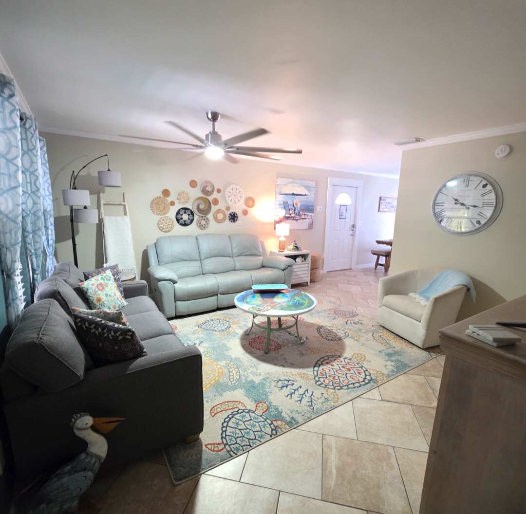 7405 Beach Dr A – Living room with gray sofas, decorative rug, wall art, lamp, and access to a front door.