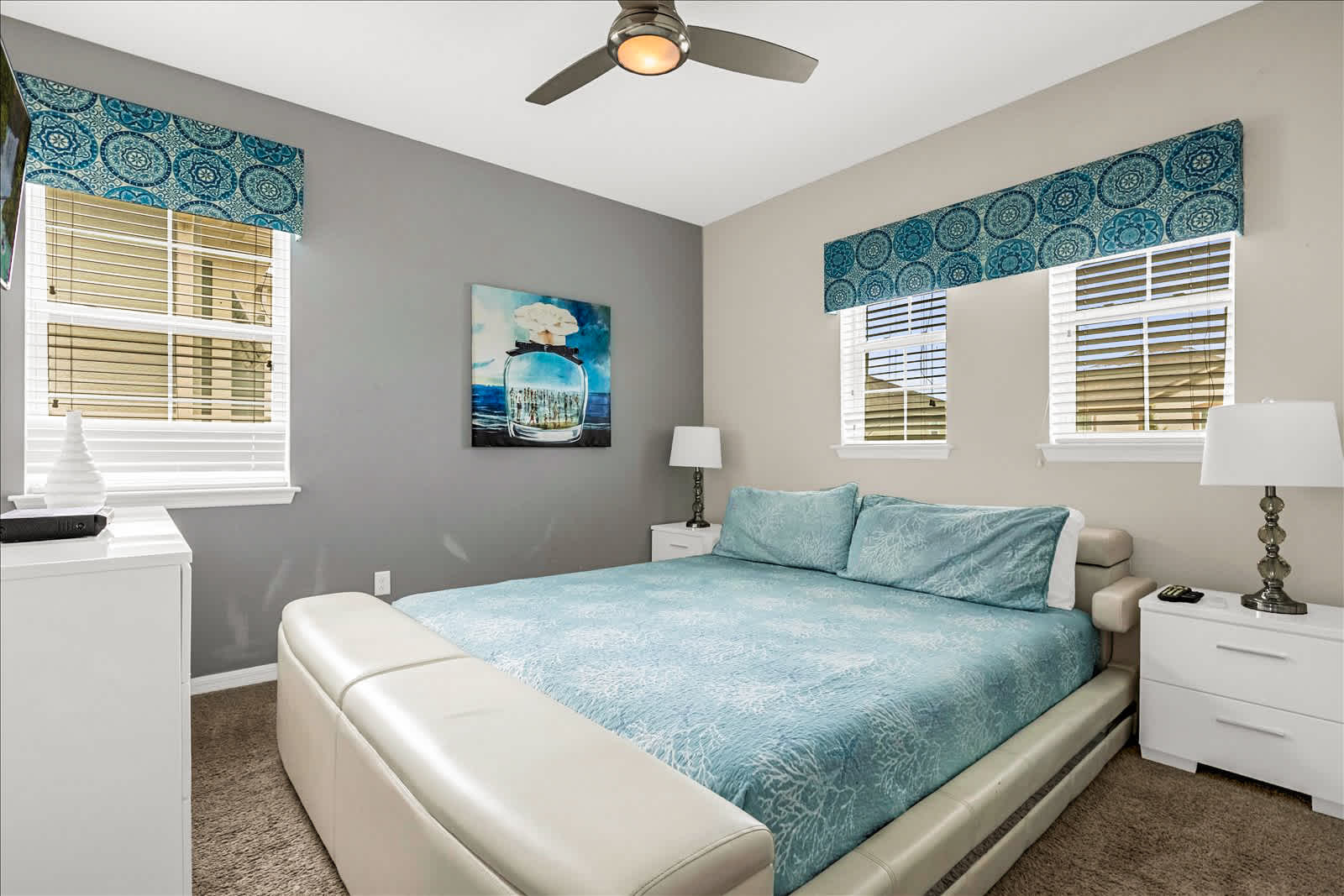 Relax in a bright bedroom with an upholstered king-size bed, aqua patterned bedding, coordinated window valances, and coastal-inspired artwork.