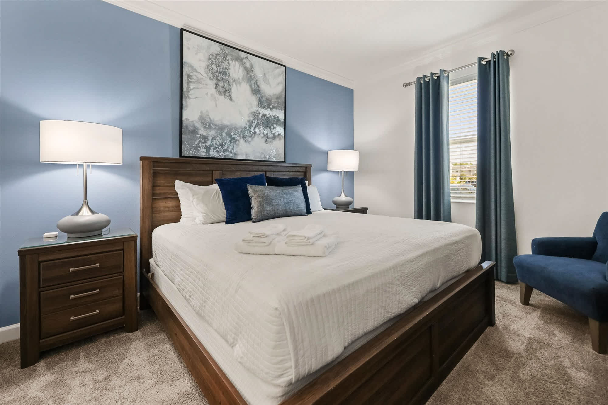 Serene bedroom with plush white bedding, navy accents, matching bedside lamps, and a blue armchair beside the sunlit window.