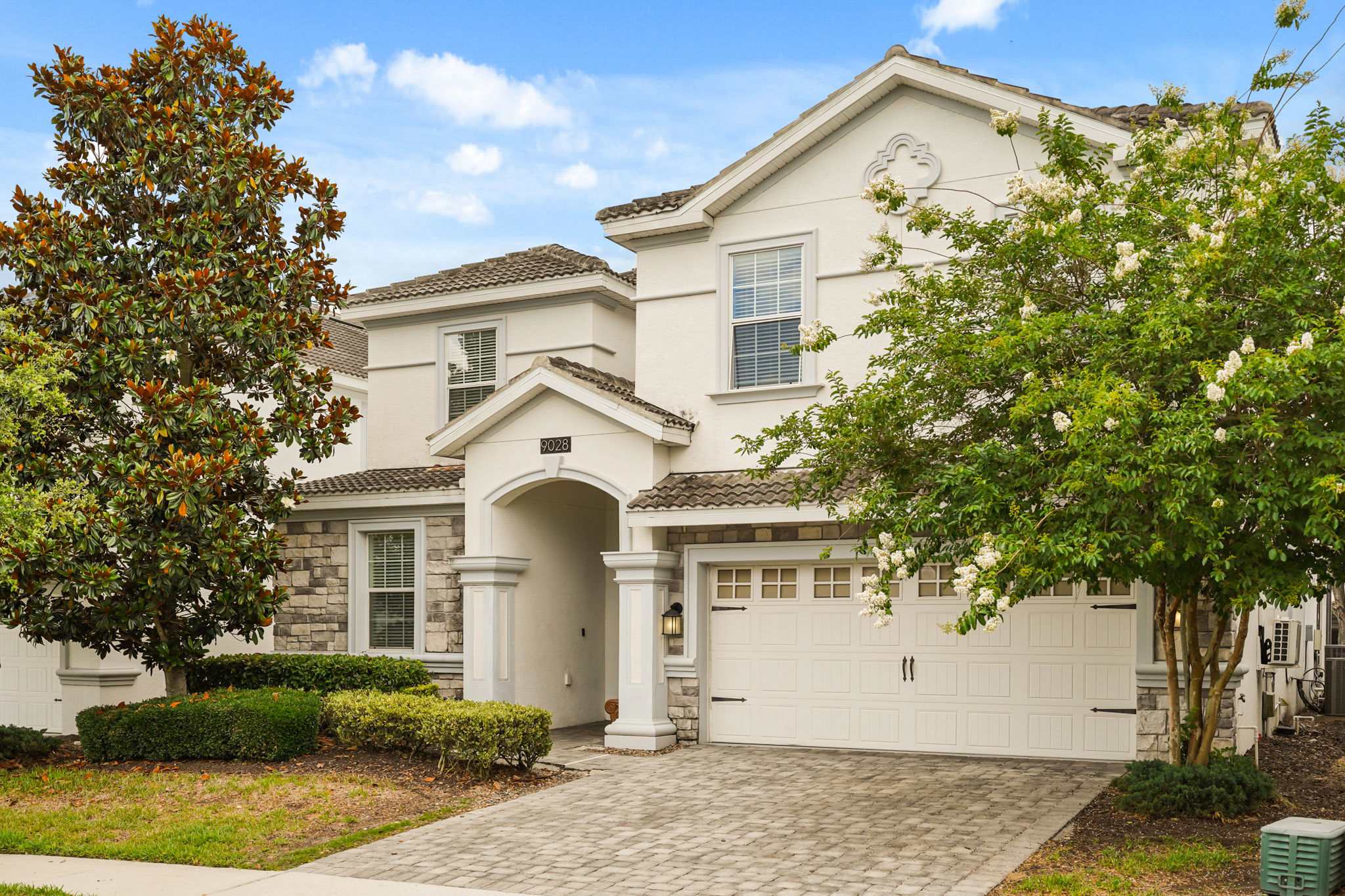 Elegant villa exterior with a covered arched entrance, stone accents, tiled roof, flowering landscaping, and a paved driveway welcoming guests.