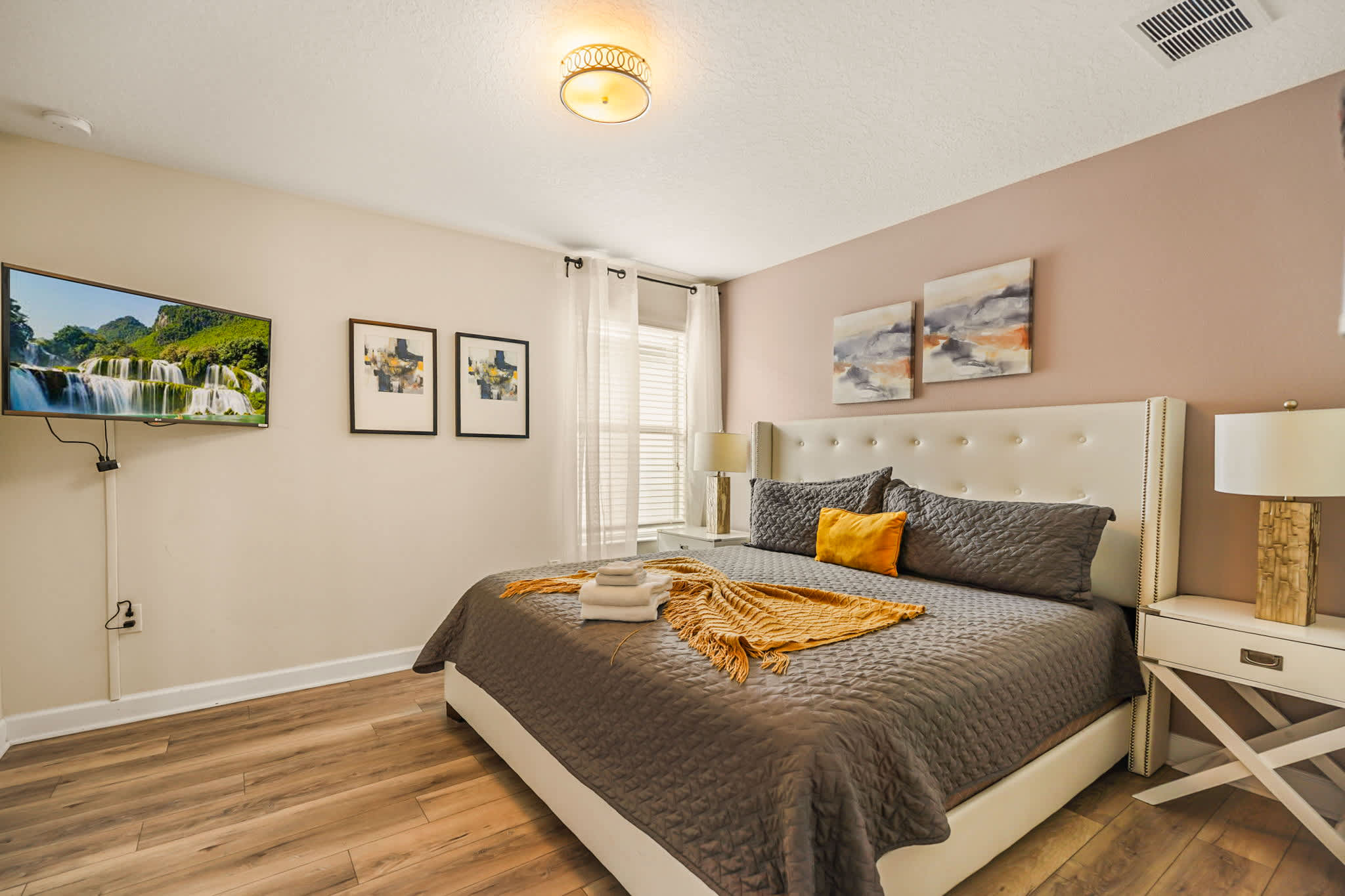 Elegant king bedroom with a tufted headboard, layered gray-and-mustard bedding, bedside lamps, and a wall-mounted TV displaying a waterfall scene.