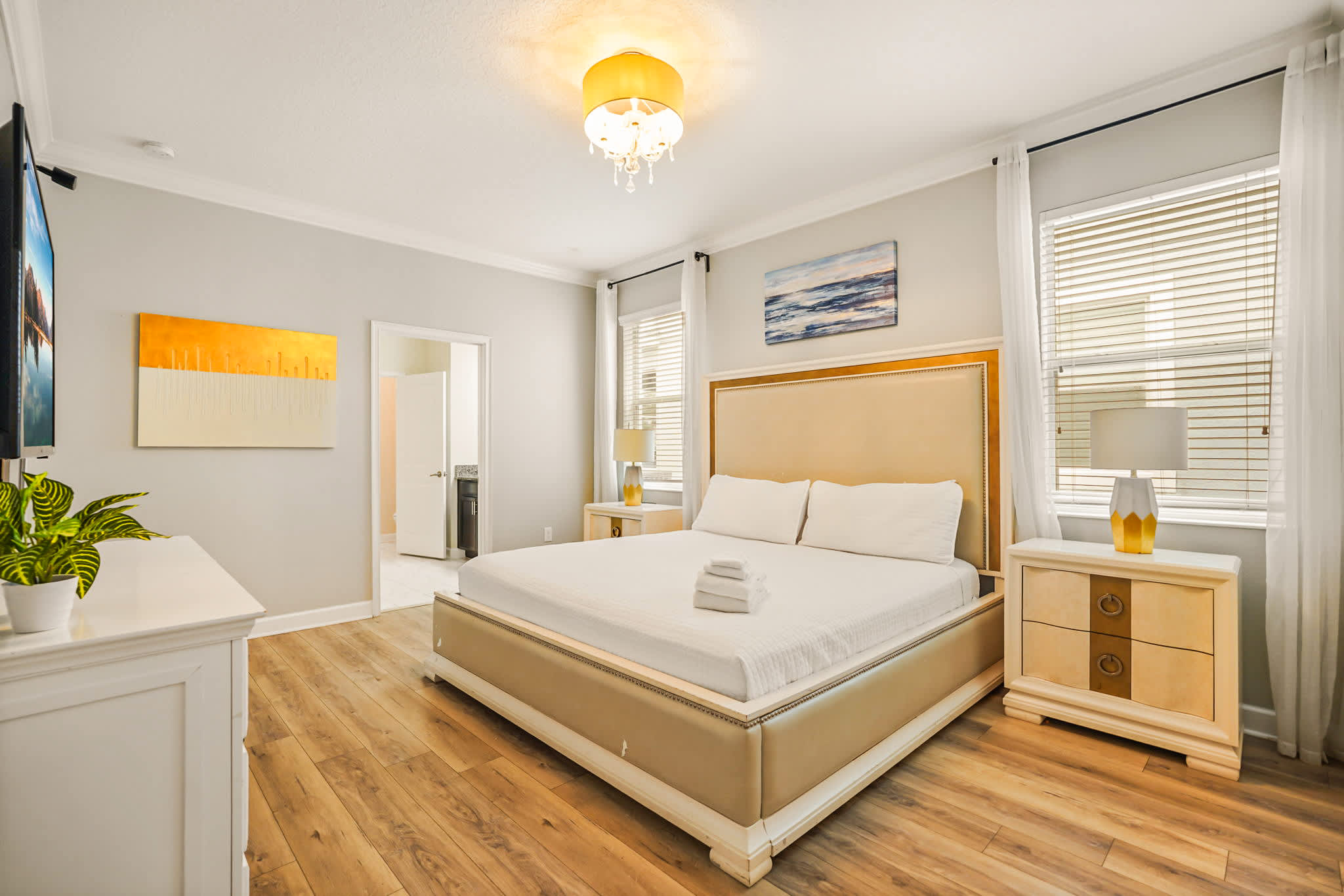 Elegant bedroom featuring a statement upholstered bed, crisp white linens, warm wood floors, and tall windows with sheer curtains and bright natural light.