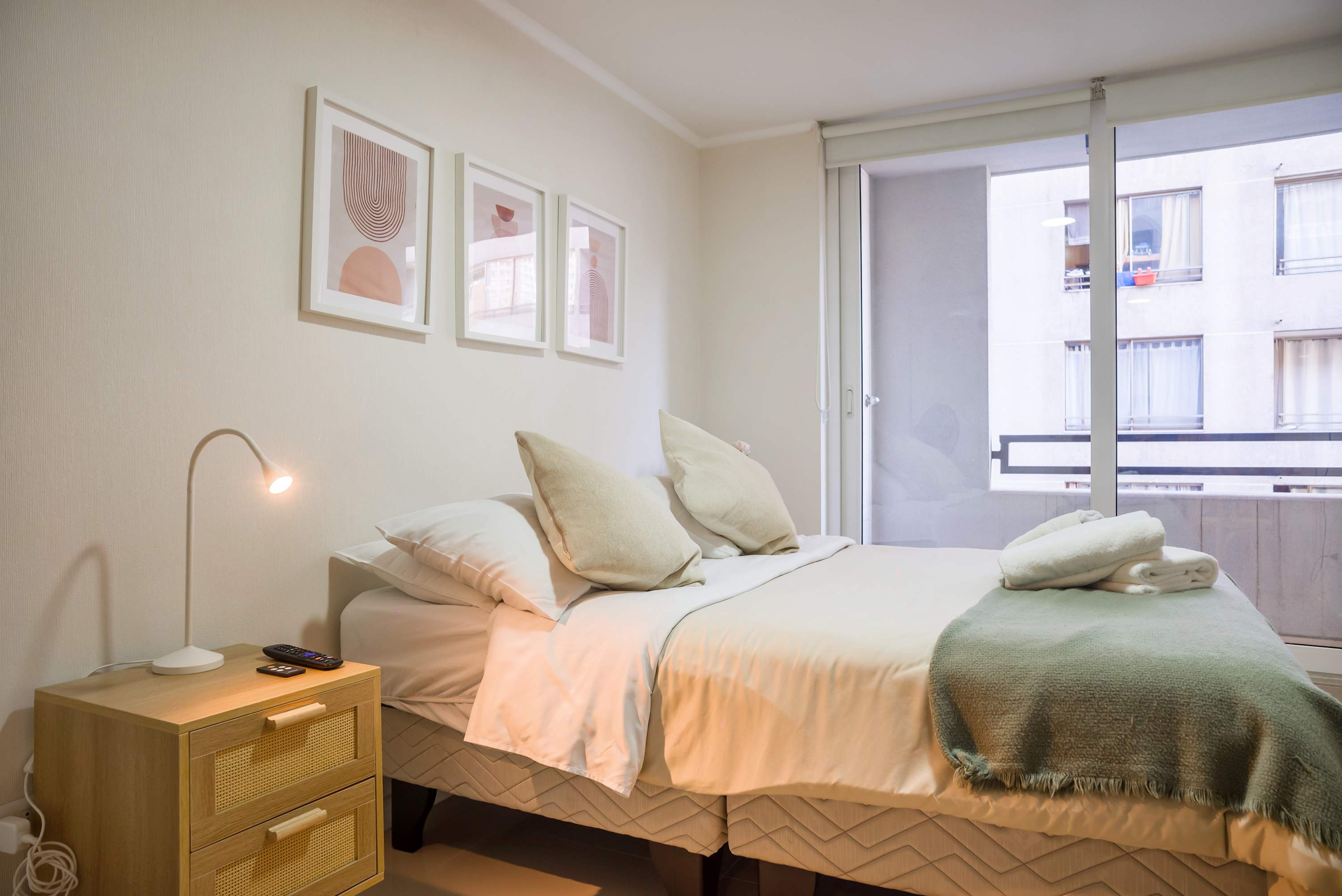 Bright studio bedroom with a comfortable double bed, soft layered bedding, framed abstract art, and a large window overlooking the neighboring city apartments.