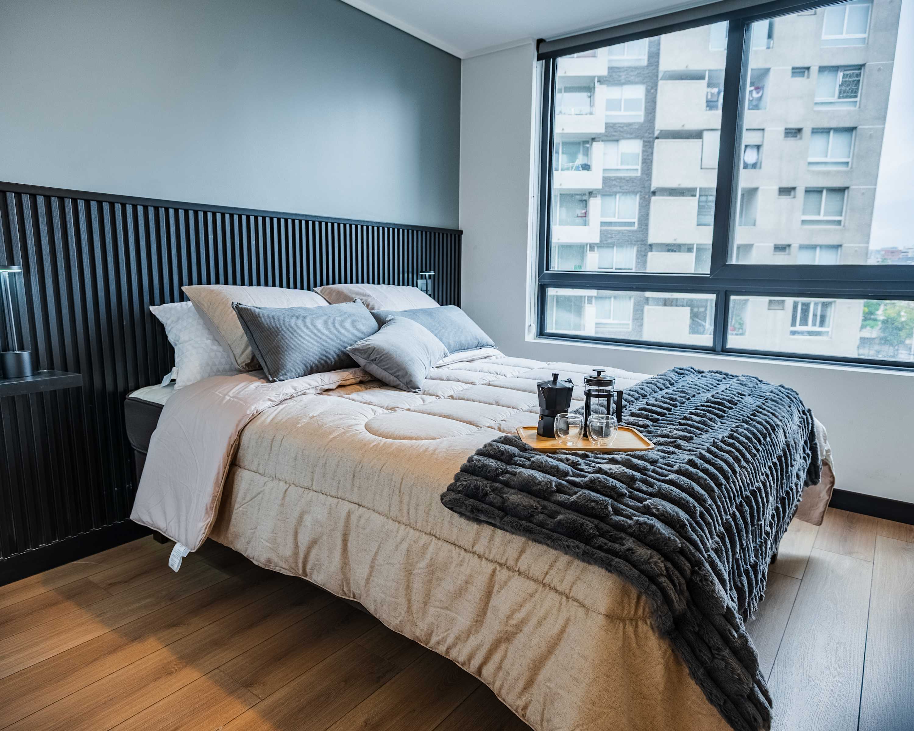 Private bedroom with a black slatted headboard, layered bedding, and a coffee tray beside large windows overlooking the surrounding city.