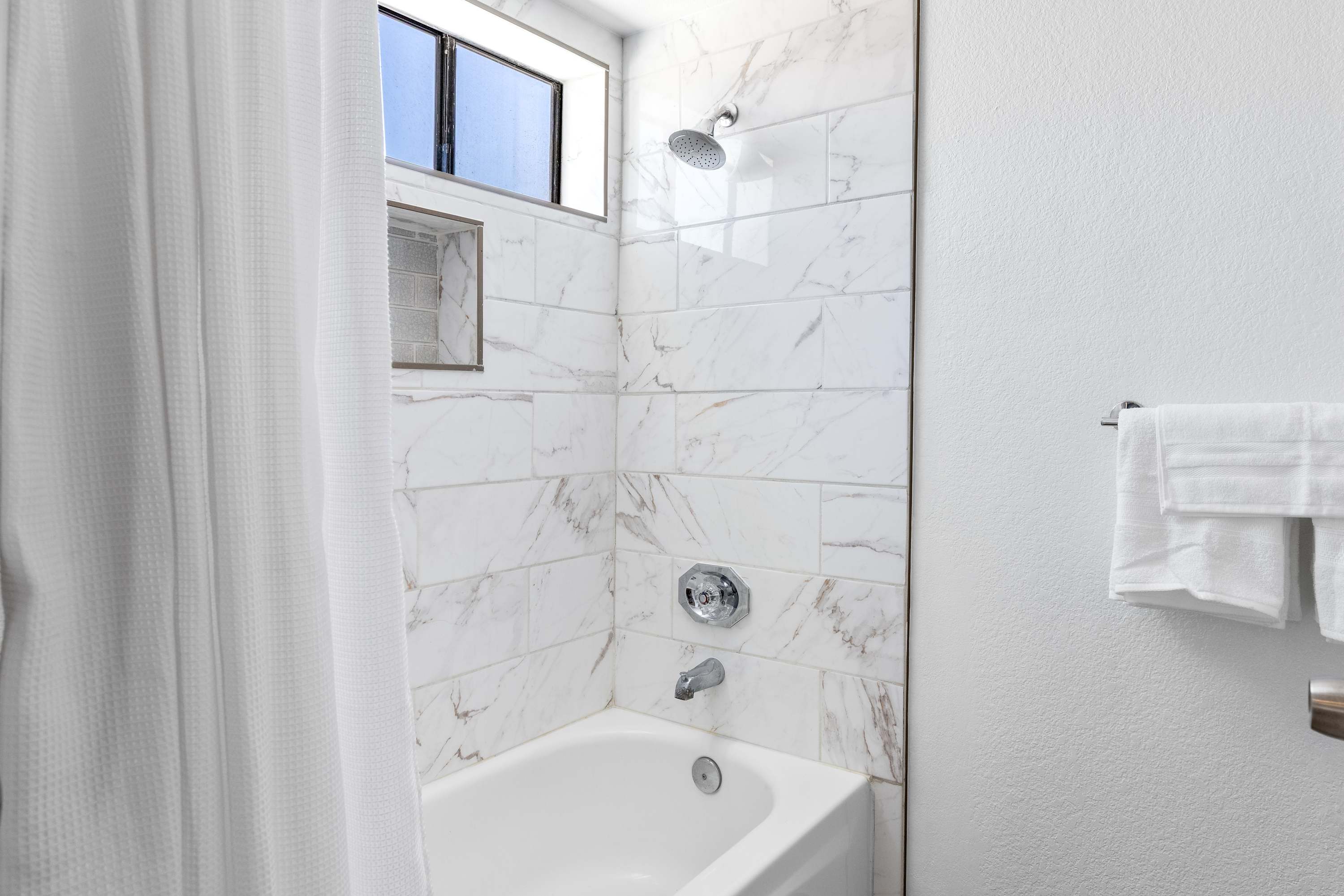 Bathroom tub and shower with marble-patterned wall tile and a high window.