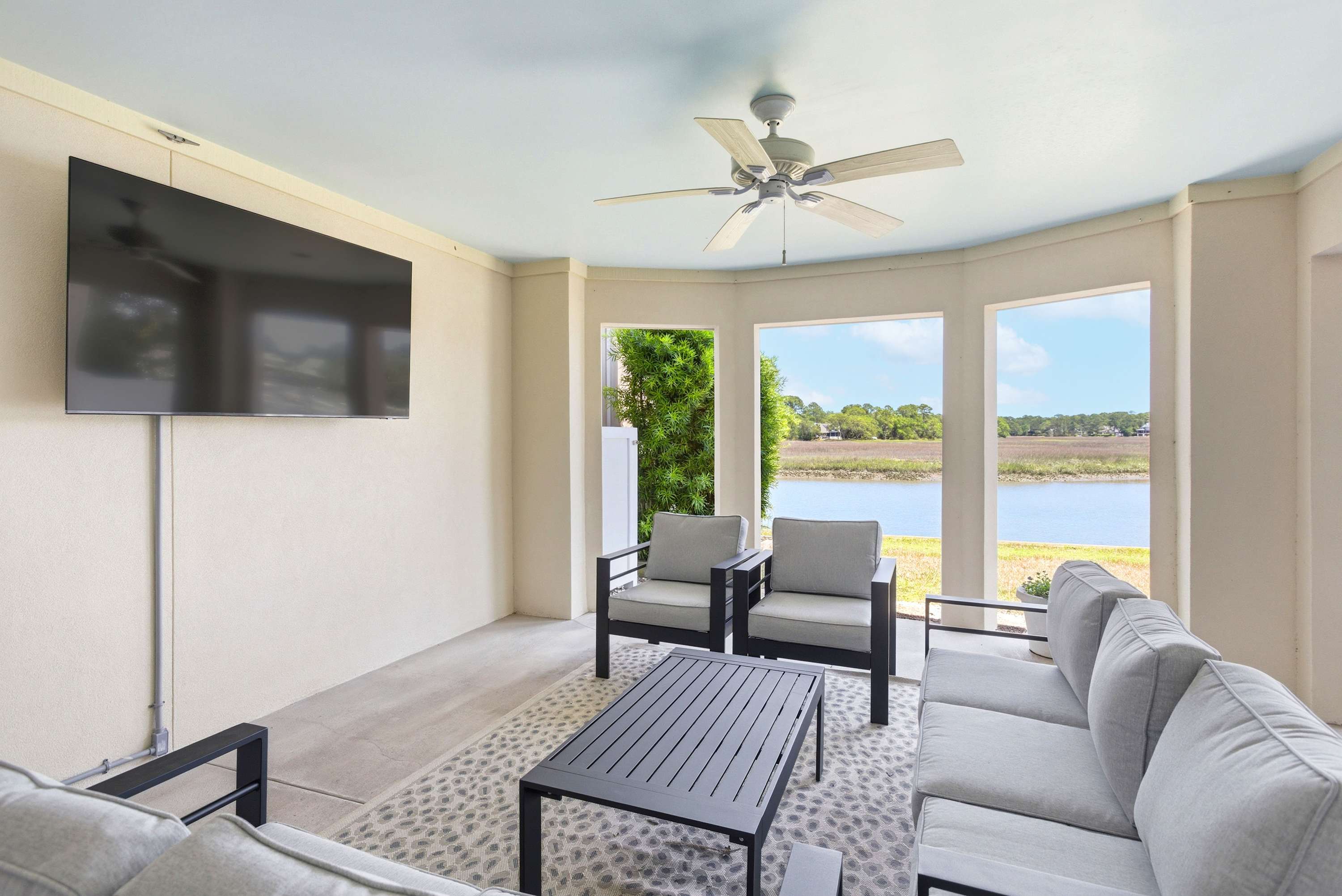Waterfront lounge with plush sectional seating, ceiling fan, and a large TV framed by open marsh views — the perfect spot for every sunset.