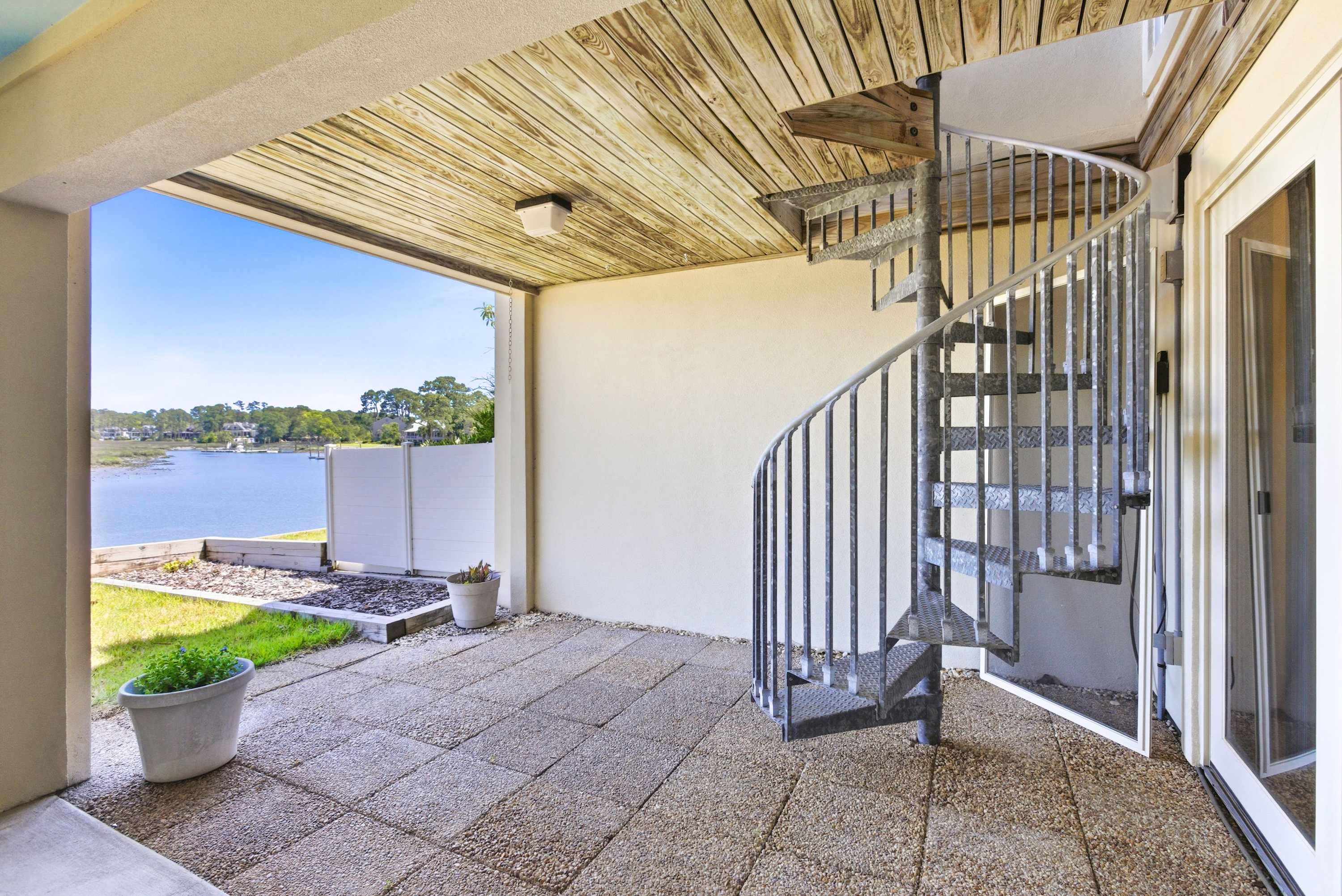 Covered ground-level terrace with wood-plank ceiling and spiral staircase leading up, all open to sweeping marsh and waterway views.