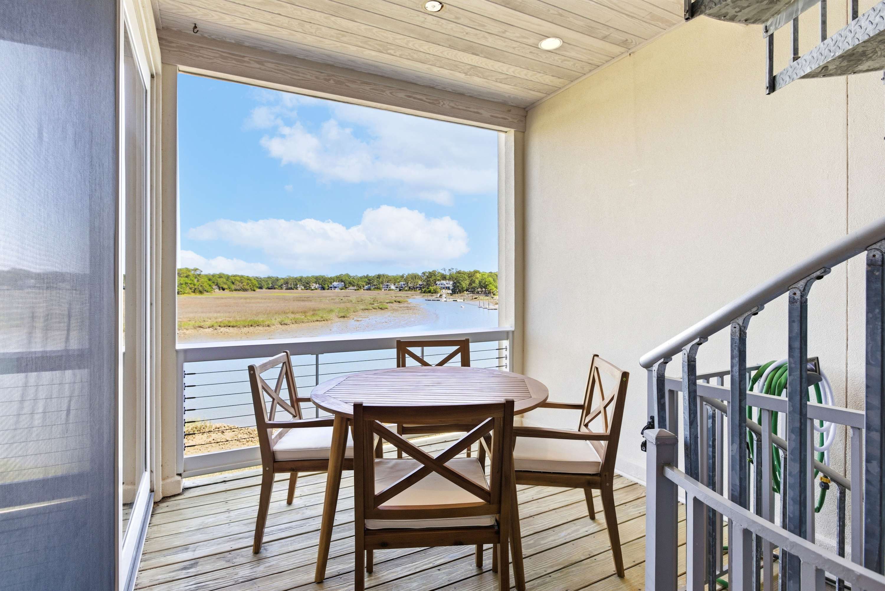 Covered mid-level deck with teak dining set and panoramic marsh and waterway views under an open blue sky.