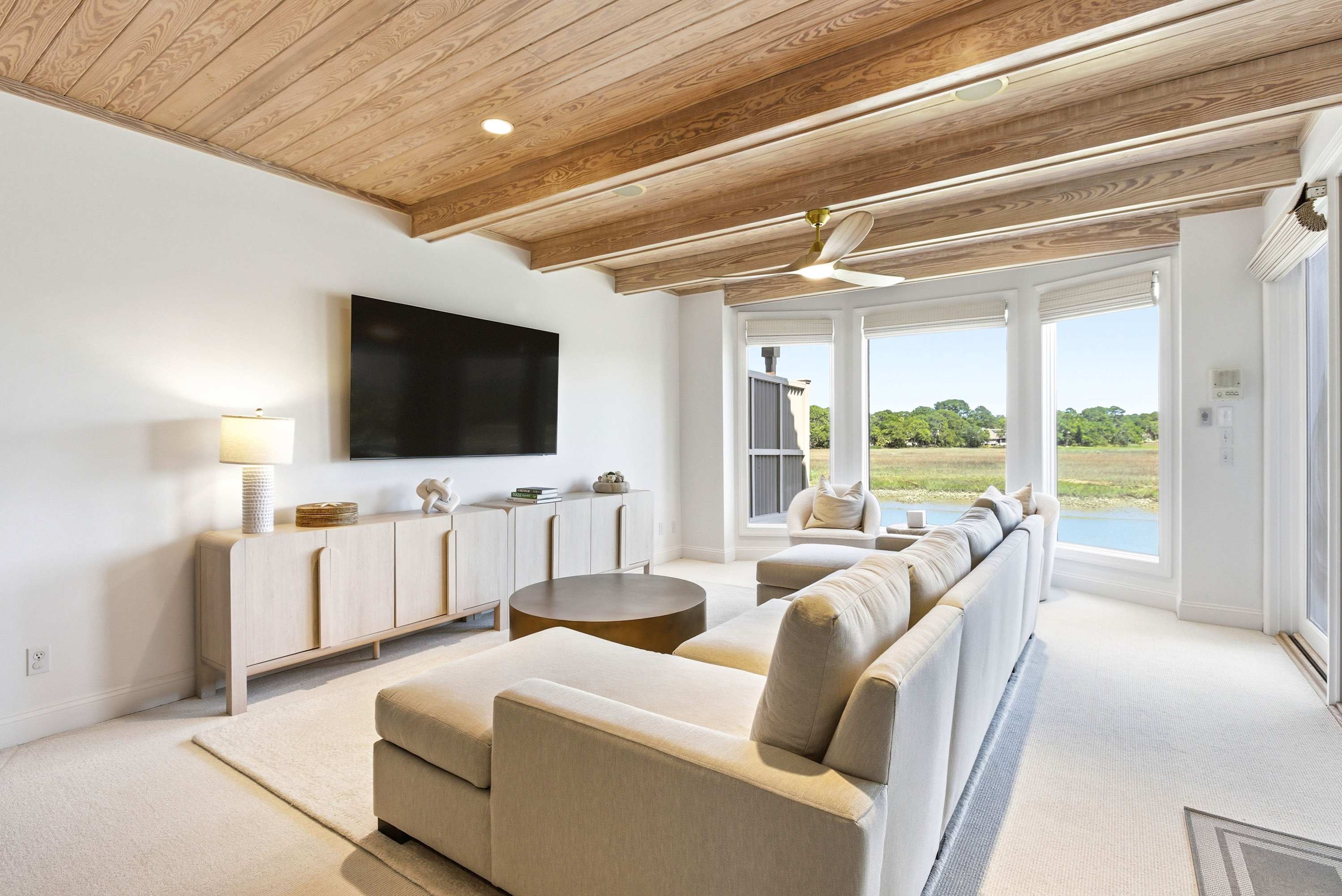 Oversized sectional, wall-mounted TV, and exposed wood-beam ceiling create the ultimate hangout — all framing a stunning waterfront backdrop.