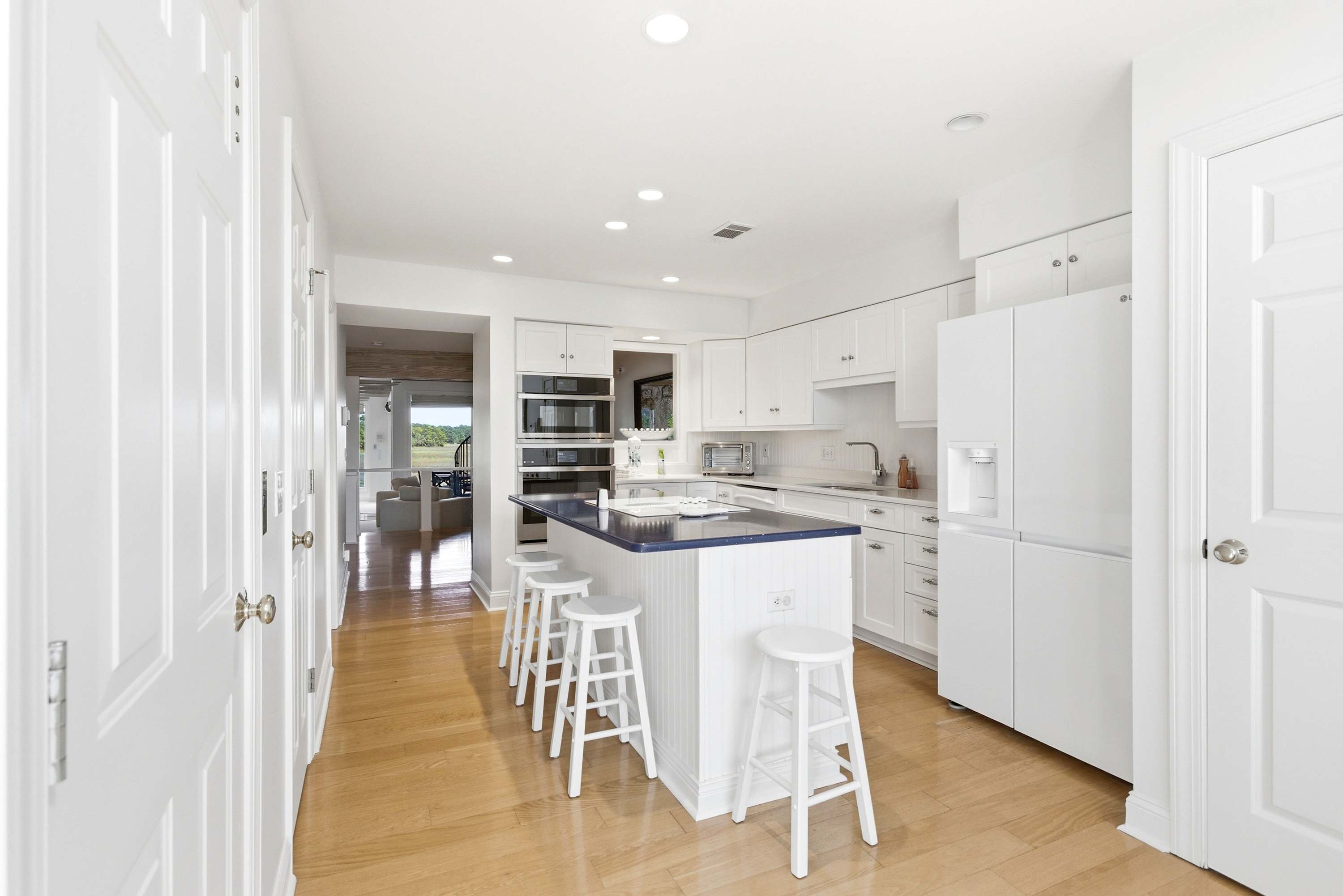 Full kitchen with double wall ovens, a large navy island with bar stools, and a hallway sightline straight to the water views beyond.