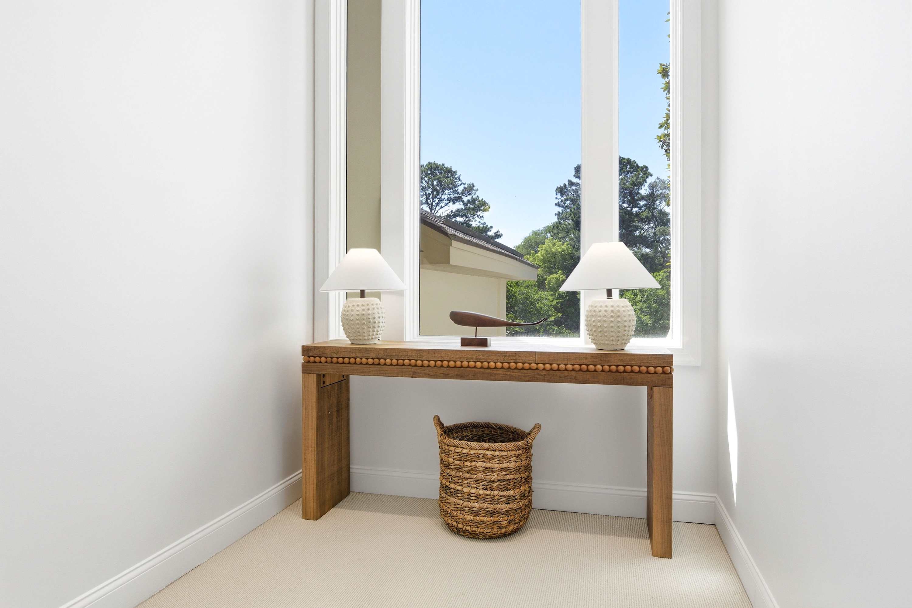 Coastal-chic console with paired ceramic lamps, a sculptural accent, and a woven basket beneath a sunlit window.