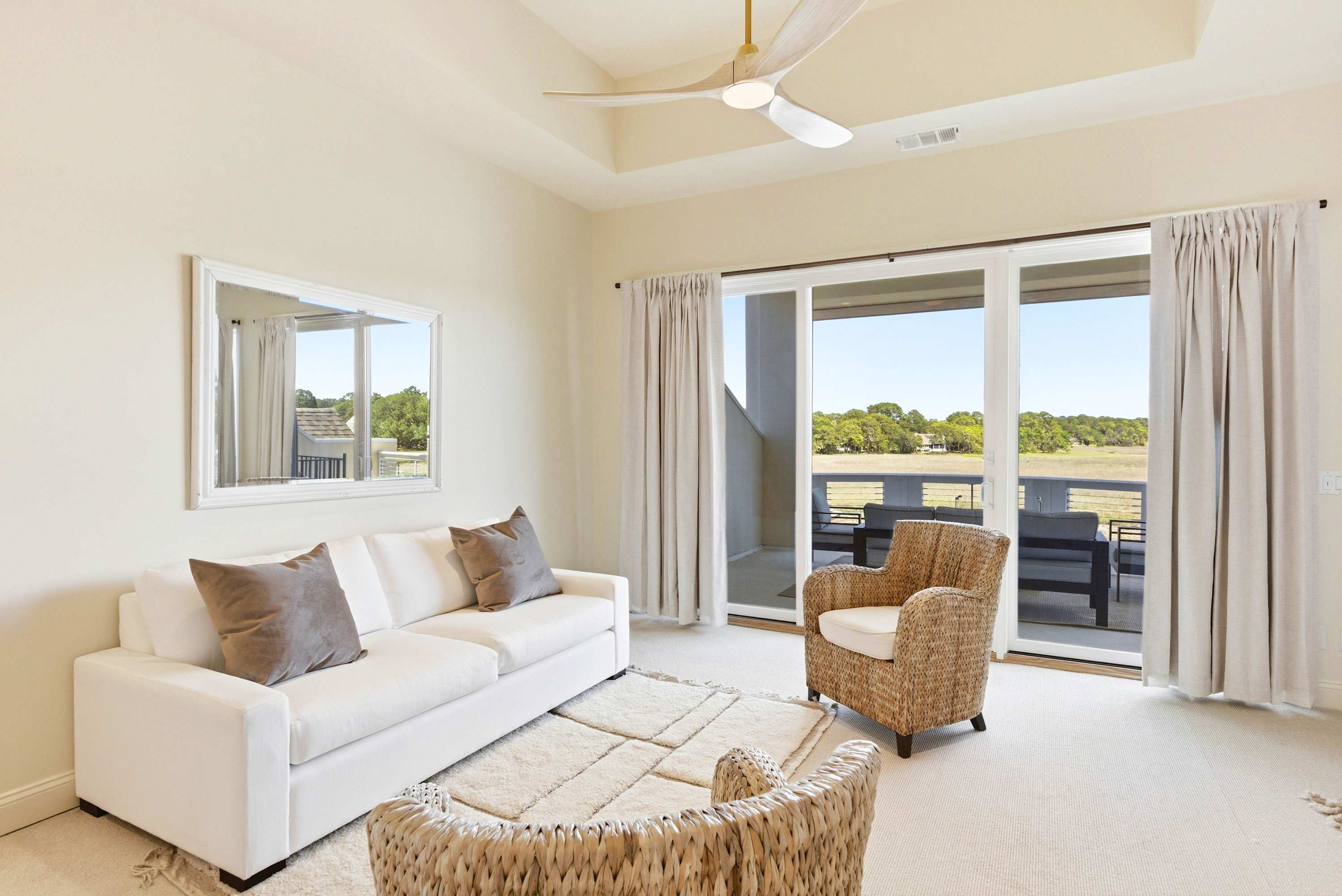 Primary suite sitting room with white sofa, woven accent chairs, linen drapes, and balcony doors opening to open marsh views.
