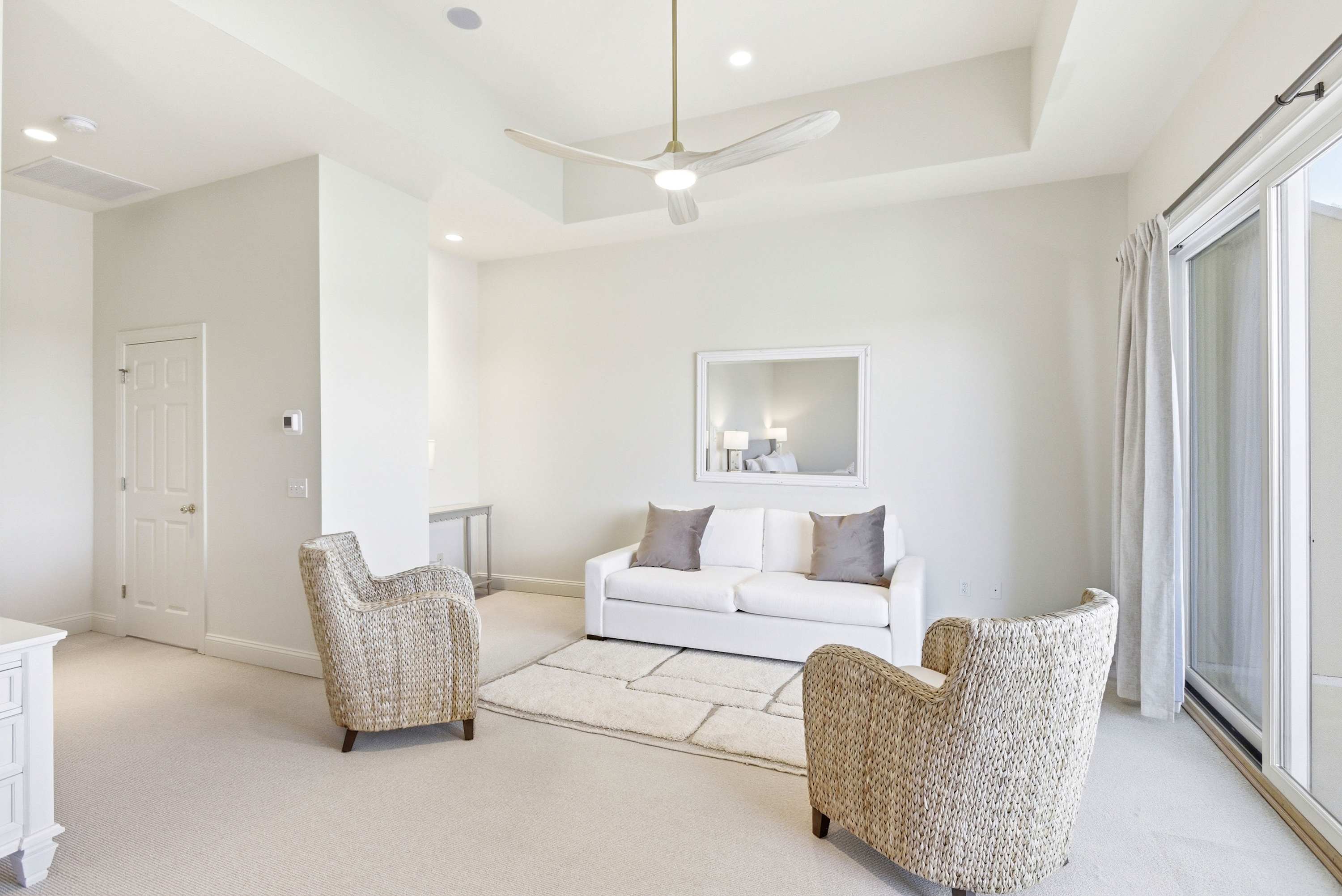 The primary suite's private lounge offers a white sofa, wicker chairs, gold ceiling fan, and sliding doors straight to the waterfront balcony.