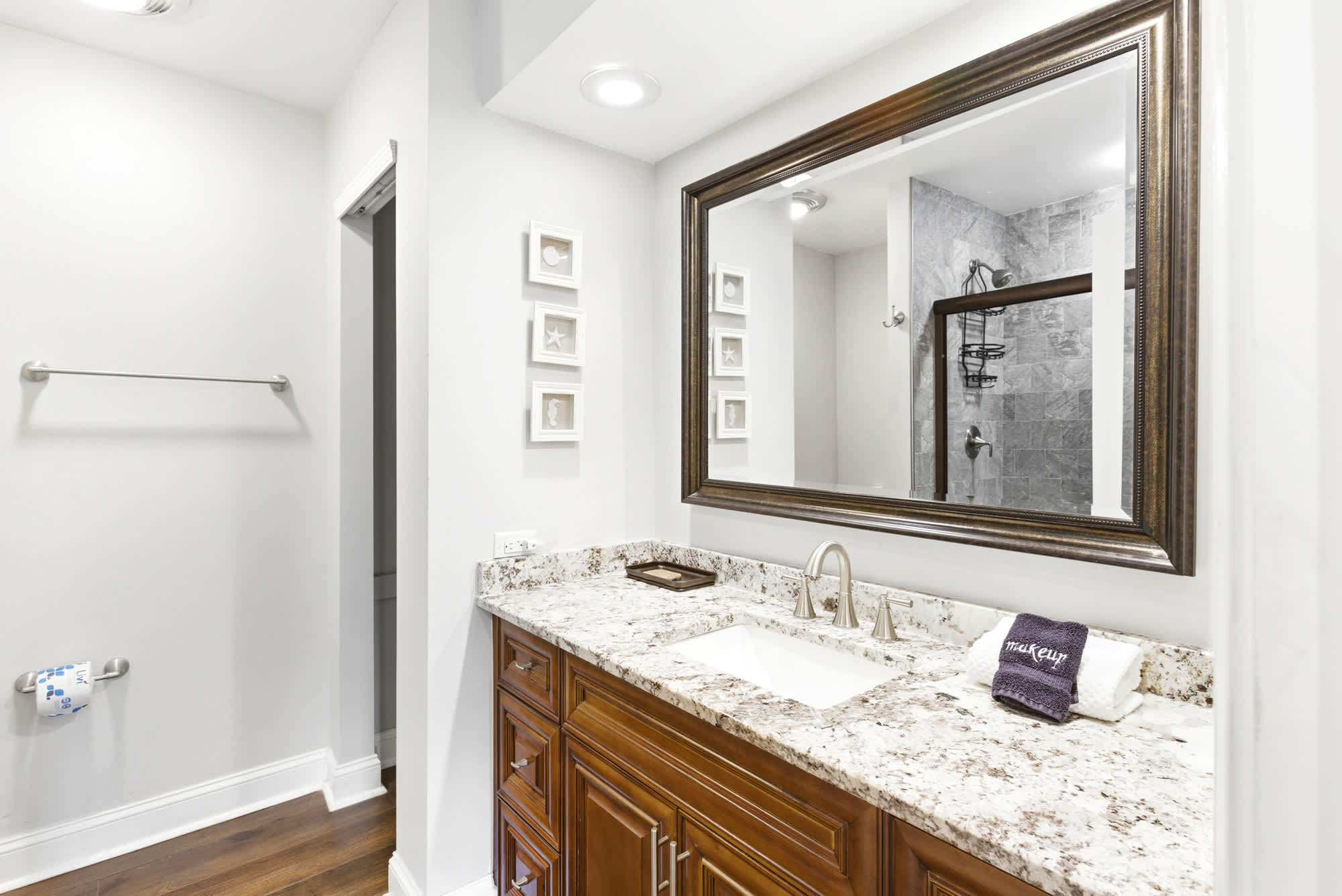 Warm wood vanity, granite countertop, oversized framed mirror, and coastal shadowbox art give this en suite a boutique hotel feel.