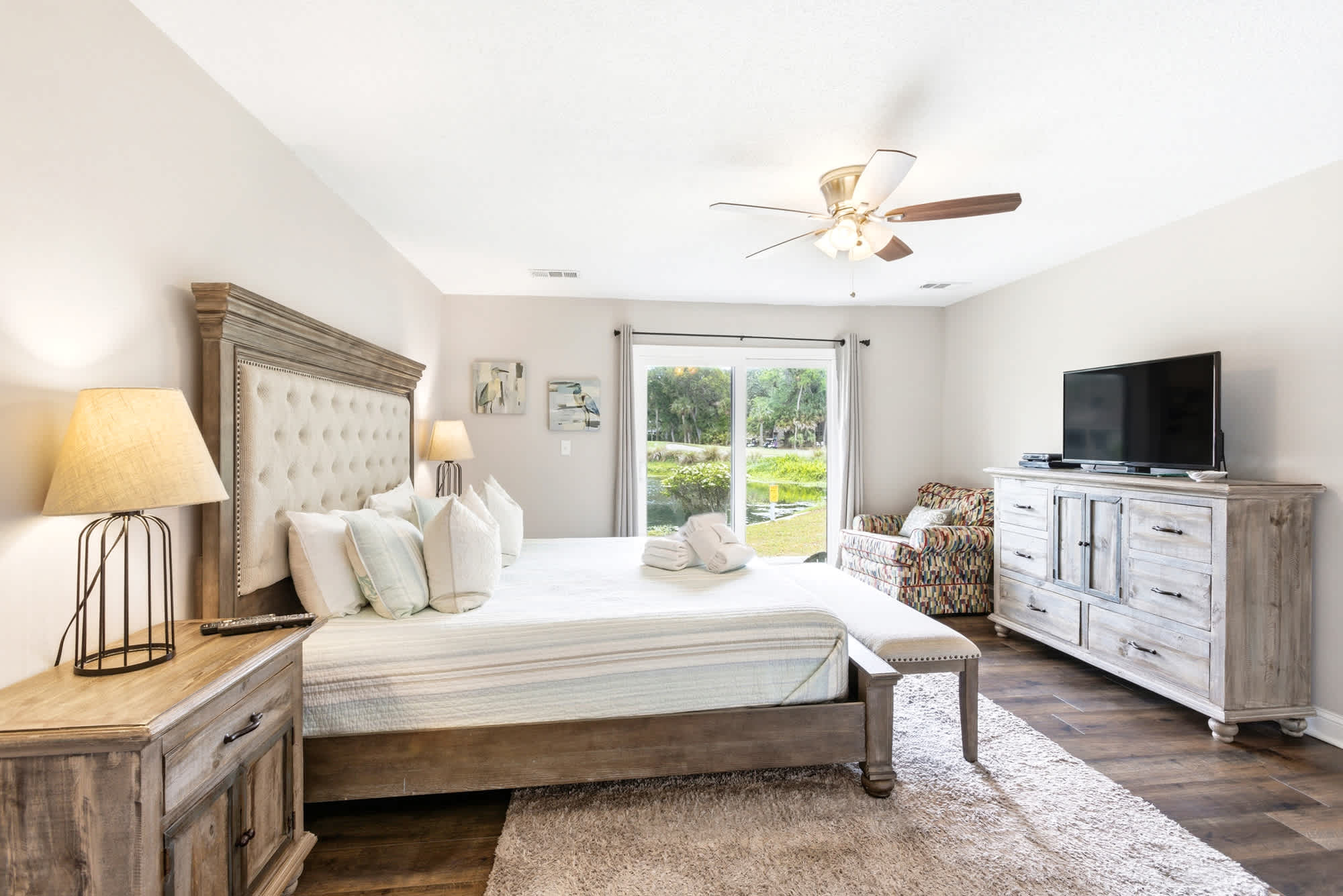 A second look at the primary suite reveals the full scope: grand headboard, TV dresser, ceiling fan, and a slider leading to the lush backyard and pond.
