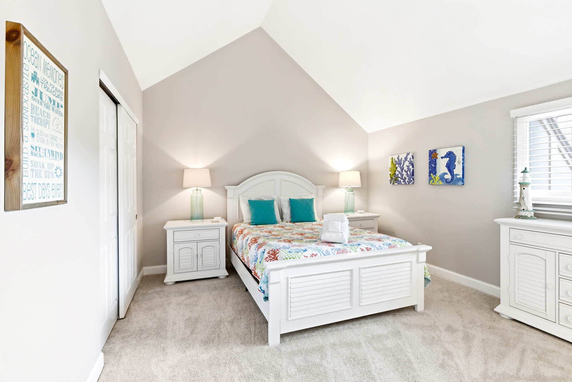 Vaulted ceiling, white cottage bed frame, a vibrant coastal quilt, and seahorse wall art make this guest room cheerful and inviting.