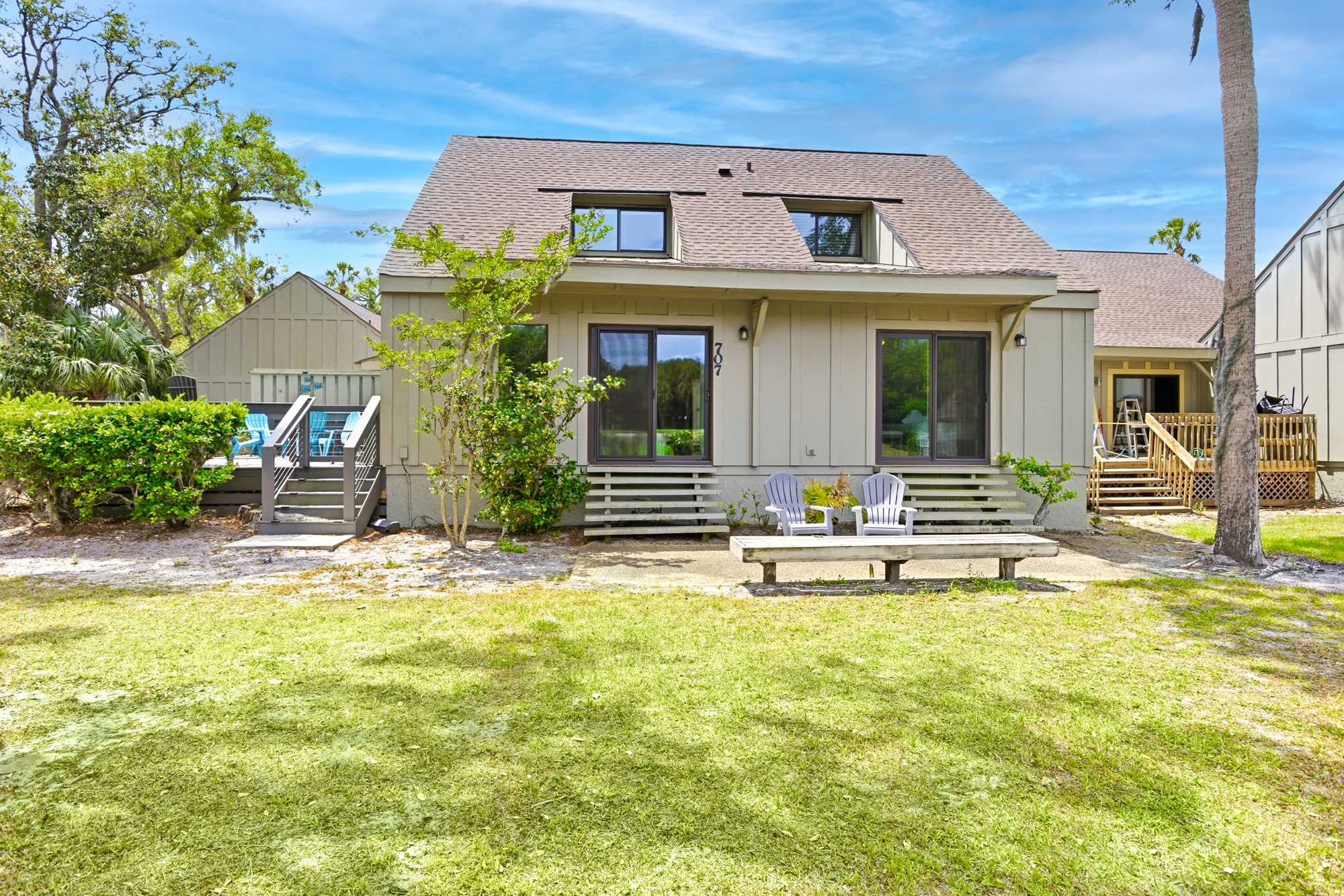 The full rear view showcases a generous green lawn, a shaded bench, and the warm, cottage-style exterior nestled among the trees.