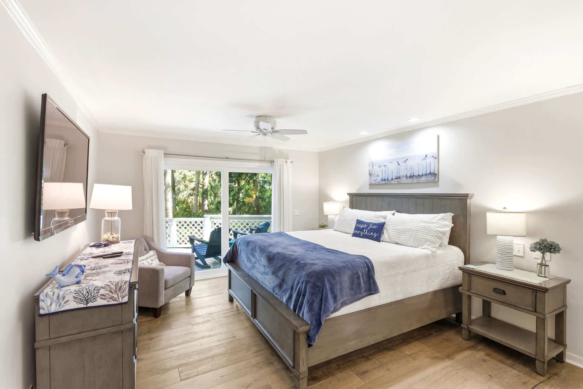 Primary king suite with a handsome gray panel headboard, indigo throw, coastal shorebird art, and sliding doors opening to a private balcony with Adirondack chairs.