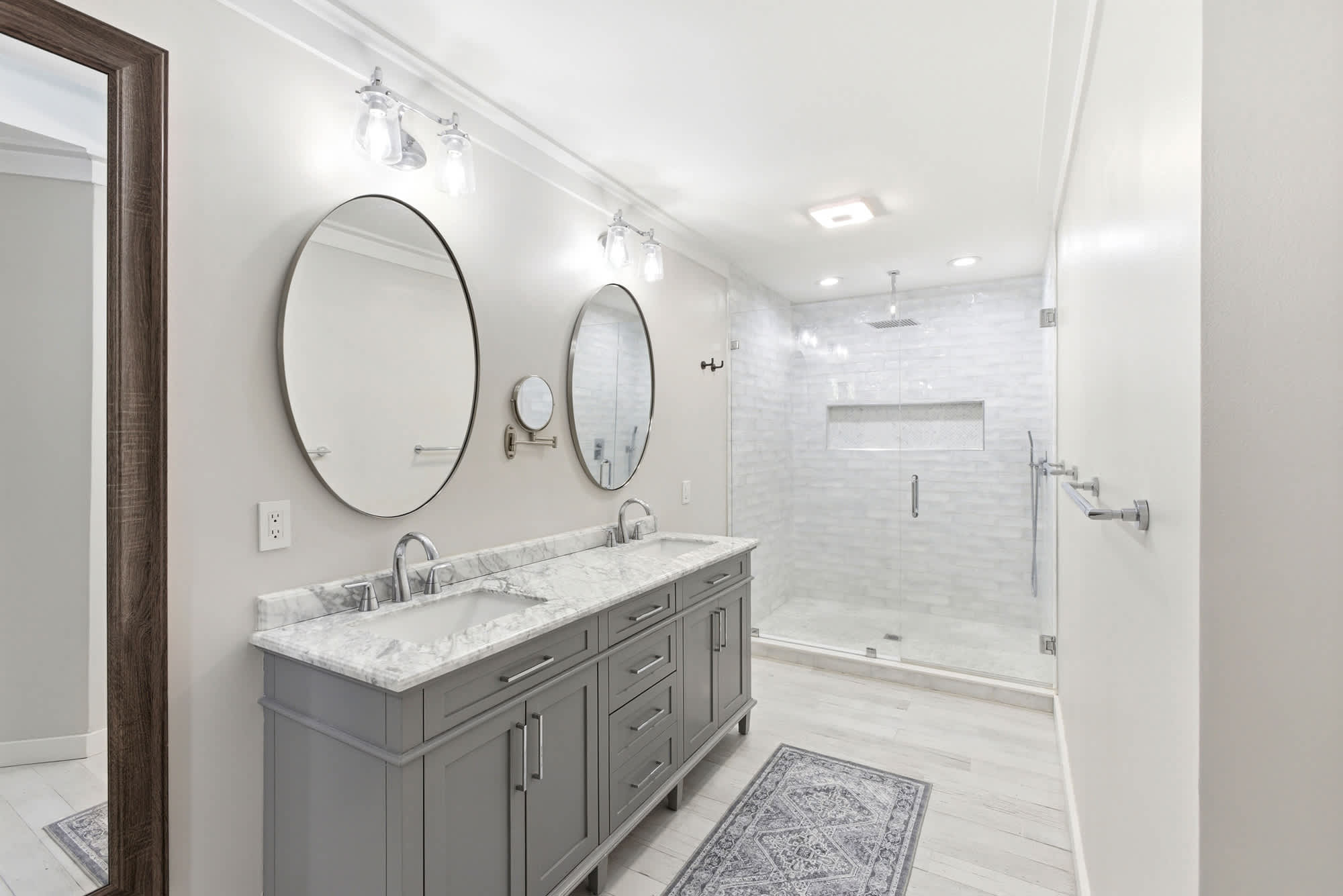 Renovated primary en suite featuring a dual gray vanity with marble countertop, twin round mirrors, and a large glass-enclosed walk-in shower with marble tile and rain shower head.