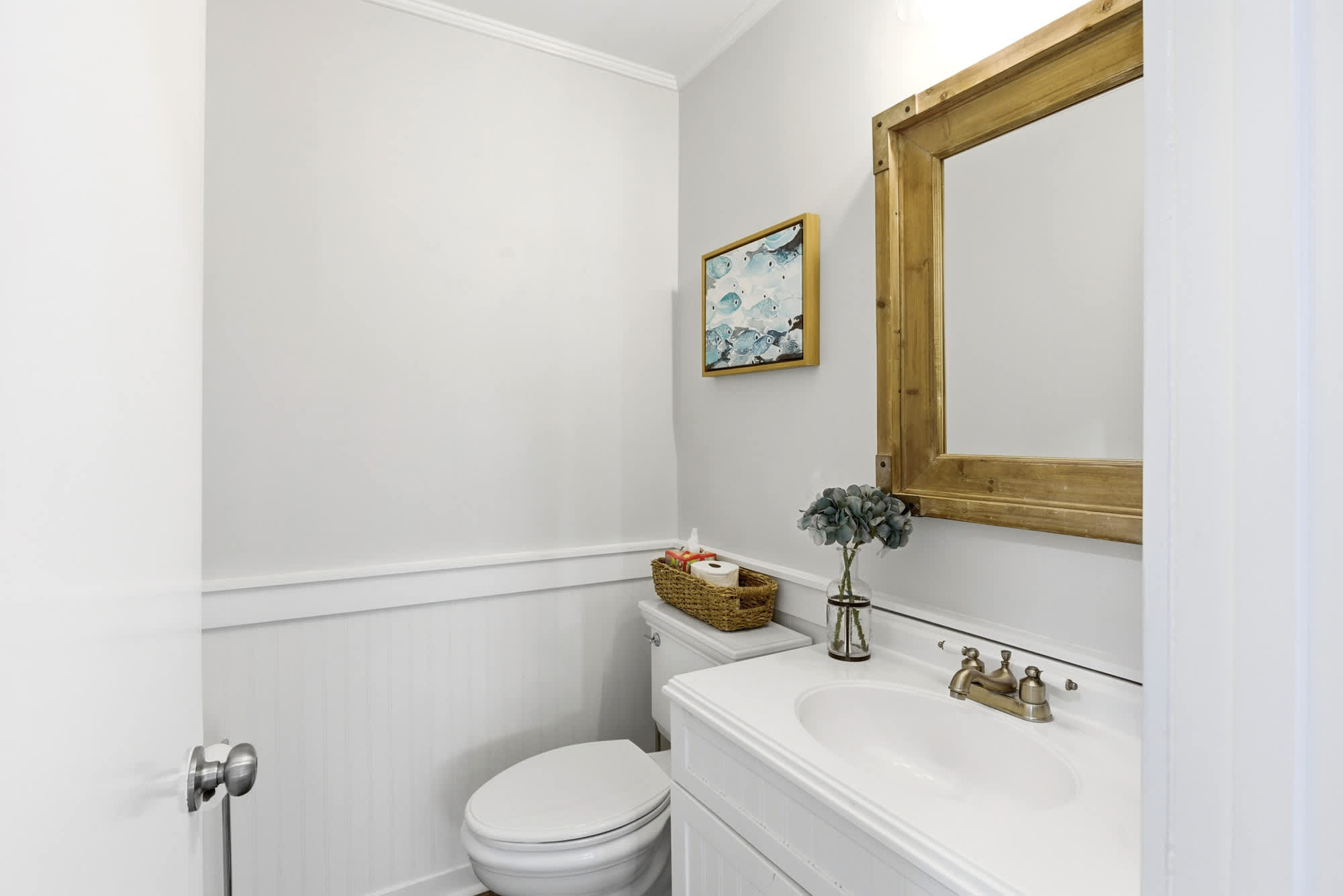 White bead-board wainscoting, a rustic gold-framed mirror, blue fish watercolor art, and a sprig of eucalyptus give this powder room a stylish, beachy personality.
