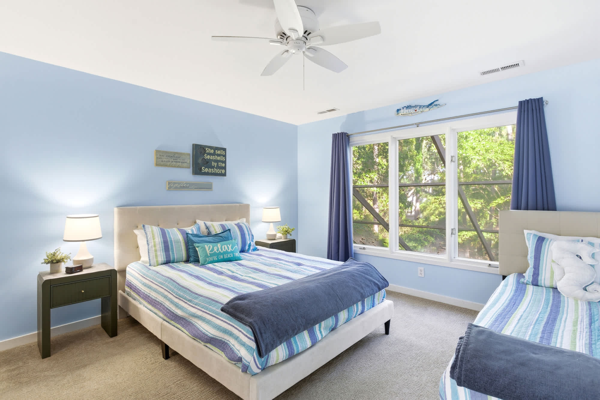 Sky-blue walls, breezy striped bedding, a 'Relax you're on beach time' pillow, and a garden window framing lush green views make this room pure coastal bliss.