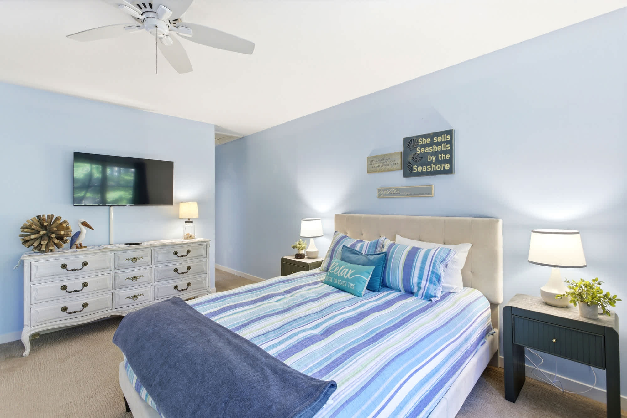 A white dresser with driftwood and pelican accents, a wall-mounted TV, and 'She sells seashells by the seashore' art add personality to this blue-walled beach room.