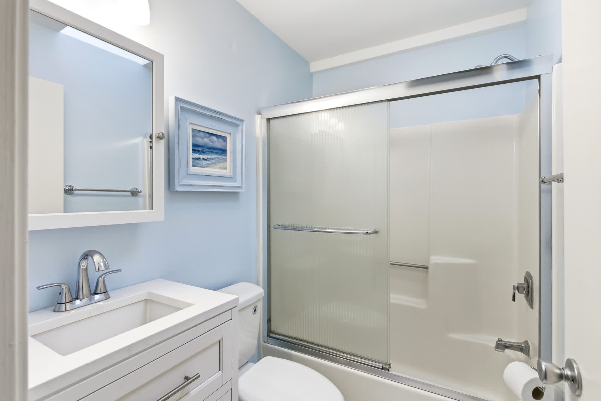 A cheerful blue bathroom with a white vanity, tub/shower combo, and framed ocean wave art — clean, bright, and perfectly on-theme.