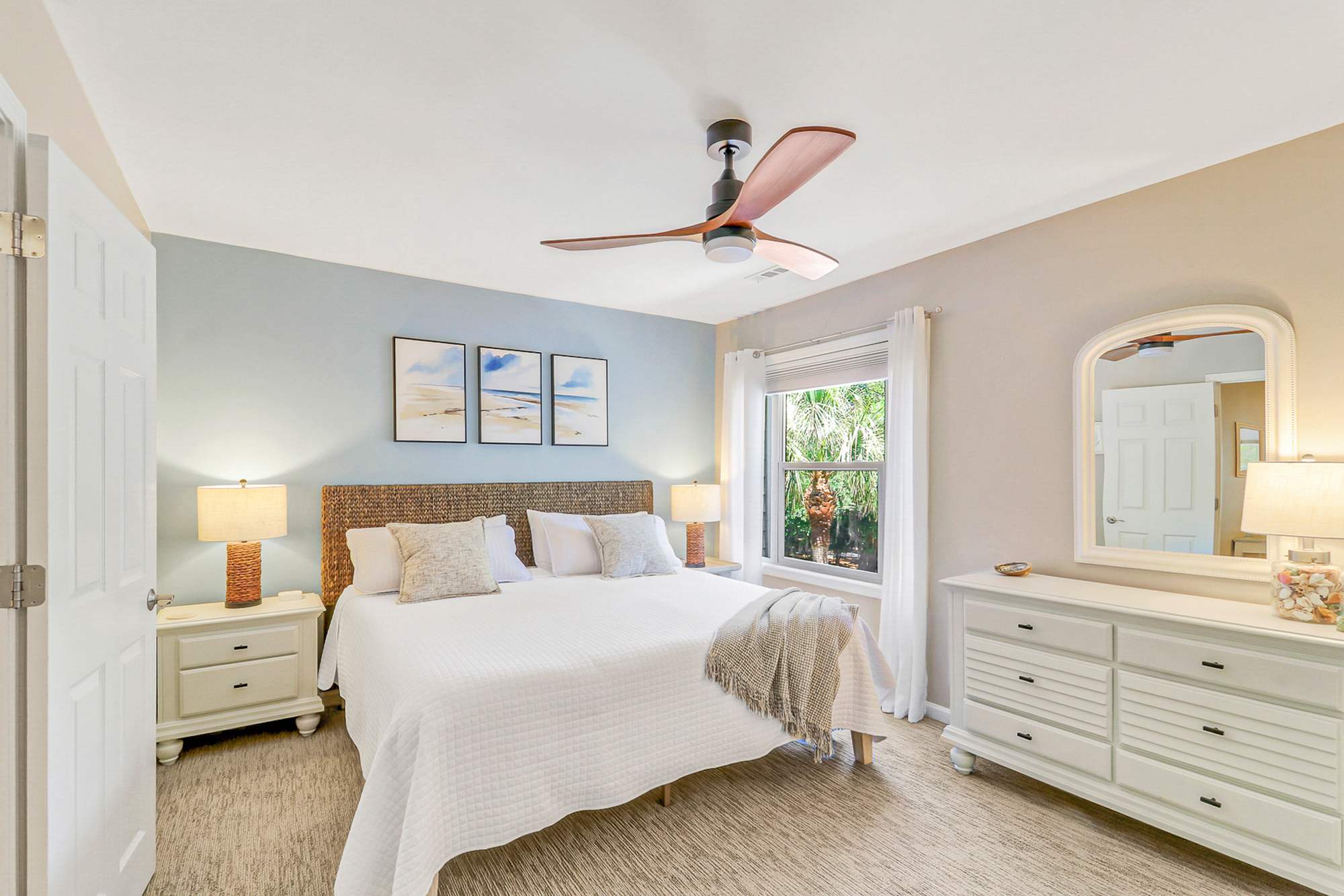 The primary bedroom feels like a boutique hotel suite with a woven rattan headboard, soft blue accent wall, beach watercolors, crisp white linens, and bedside lamps. En-suite bath included.