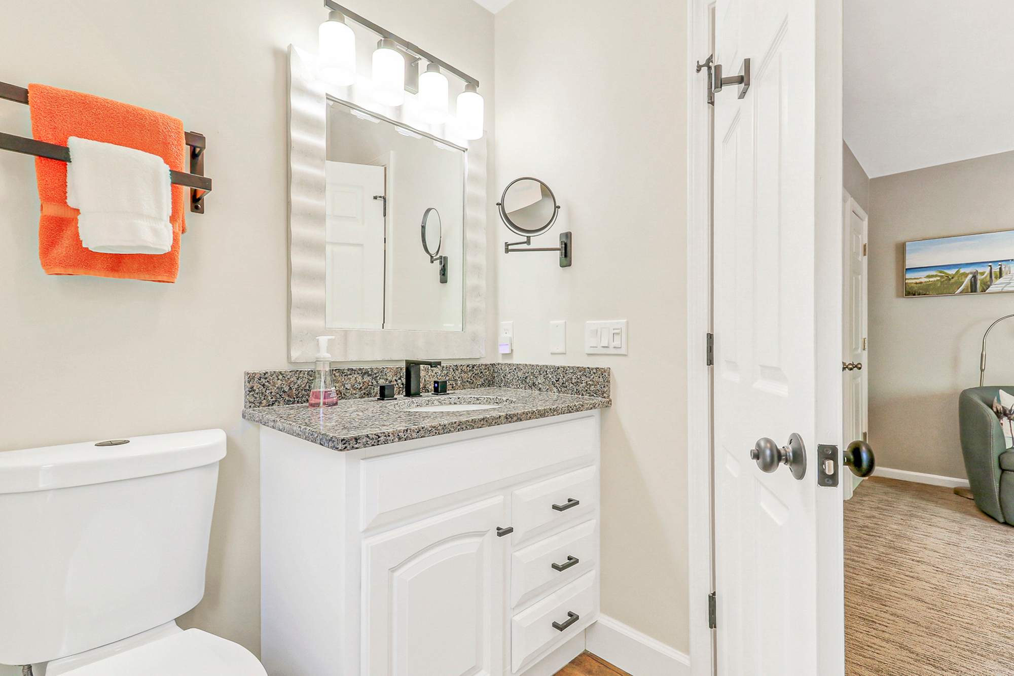 Private en-suite vanity with white cabinet, granite top, lighted mirror, and swing-arm magnifying mirror. Everything guests need to get ready comfortably, steps from the king bed.