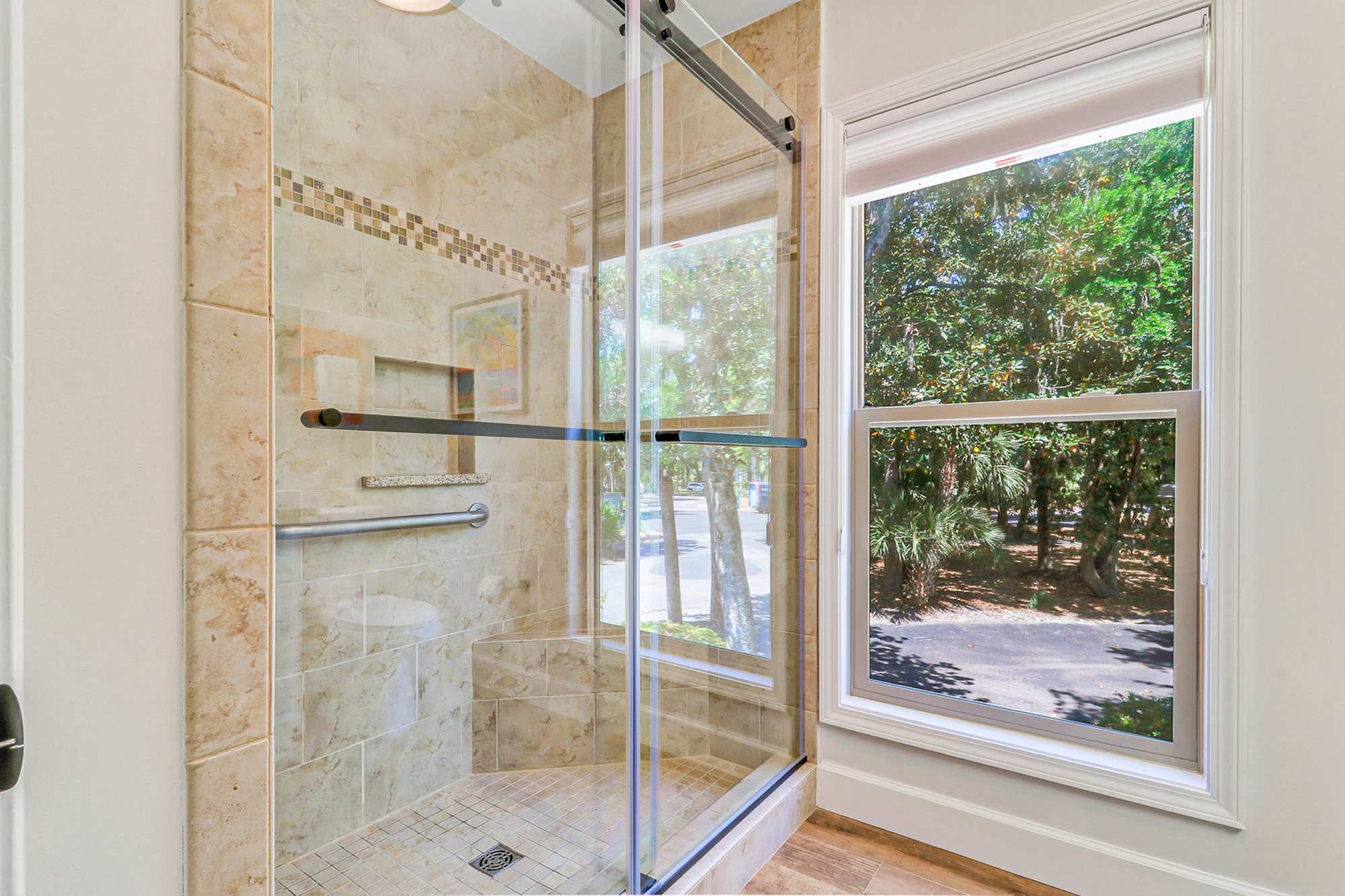 Spacious walk-in shower with travertine-style tile, built-in shelf, grab bar, and a frameless glass door. Natural light pours in from the large window beside the shower.