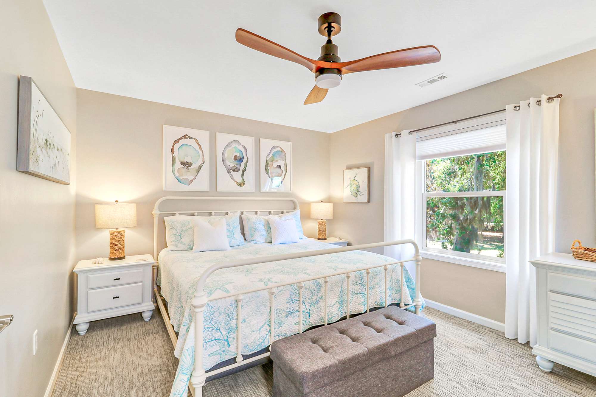 The second bedroom is a bright, cozy retreat with a white iron bed, coral-print bedding, oyster shell art, and a garden view. White furniture, a tufted ottoman, and good storage for guest luggage.