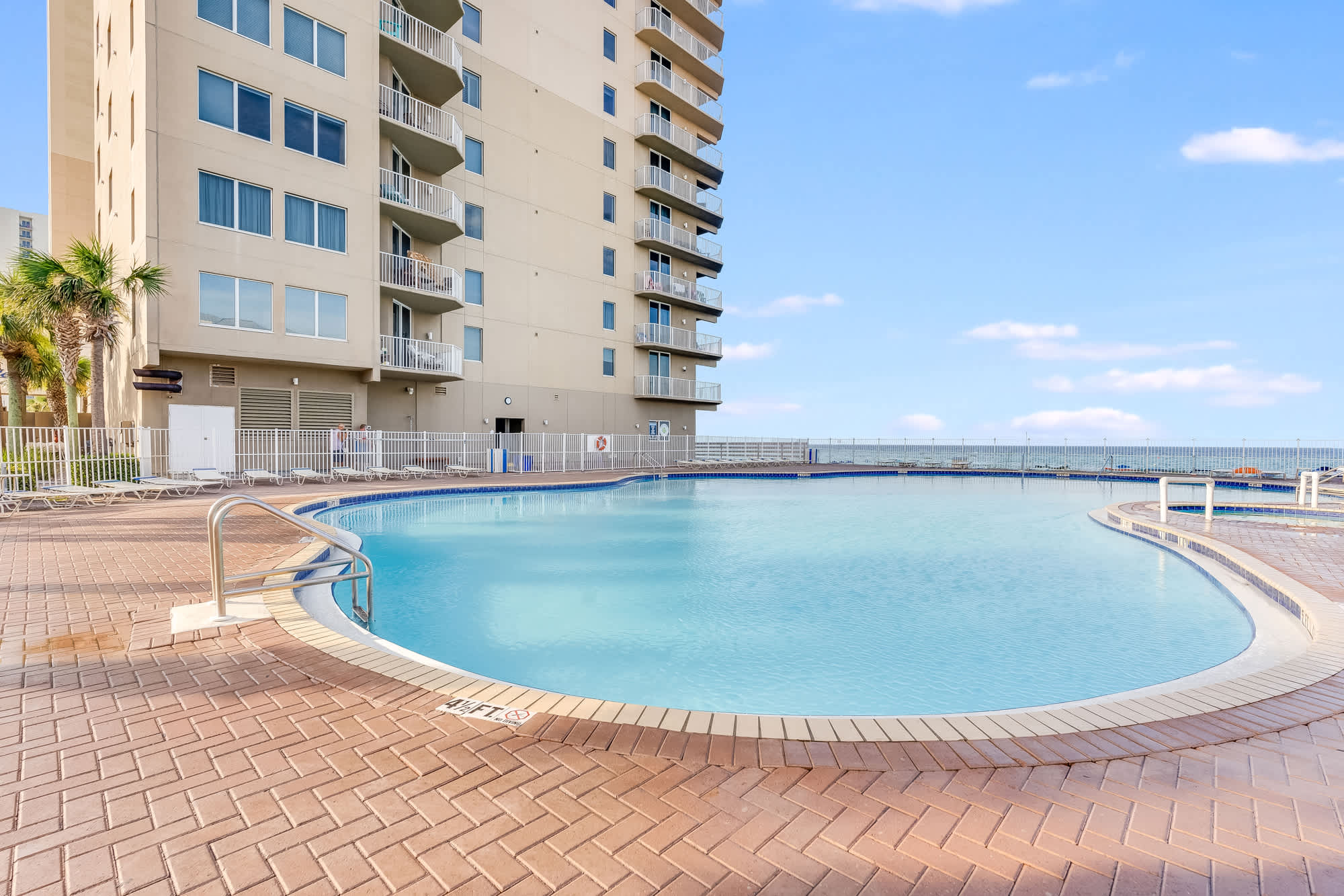 Tidewater 200 – Pool area with curved edge, tan brick deck, and building with balconies in the background.