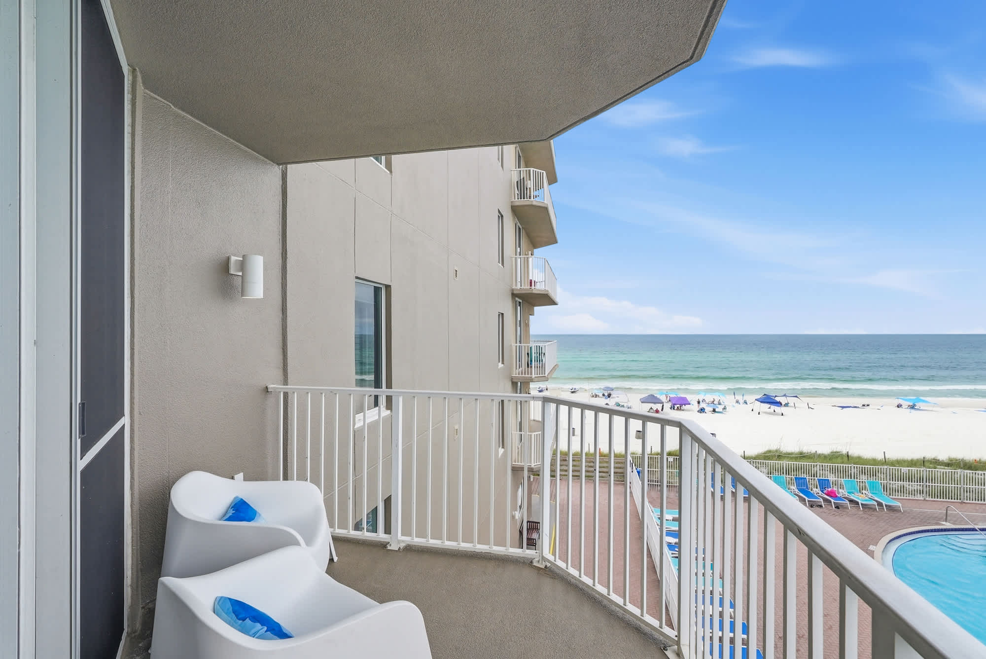 Tidewater 200 – Balcony view with two white chairs, overlooking a pool and beach with umbrellas and waves in the background.