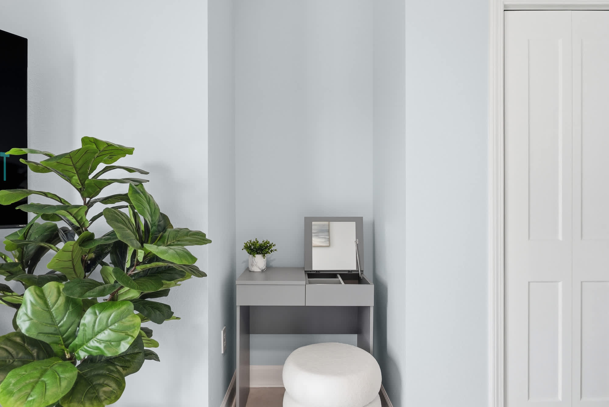 Tidewater 200 – Small corner vanity with mirror, potted plants, and white stool against light blue wall.