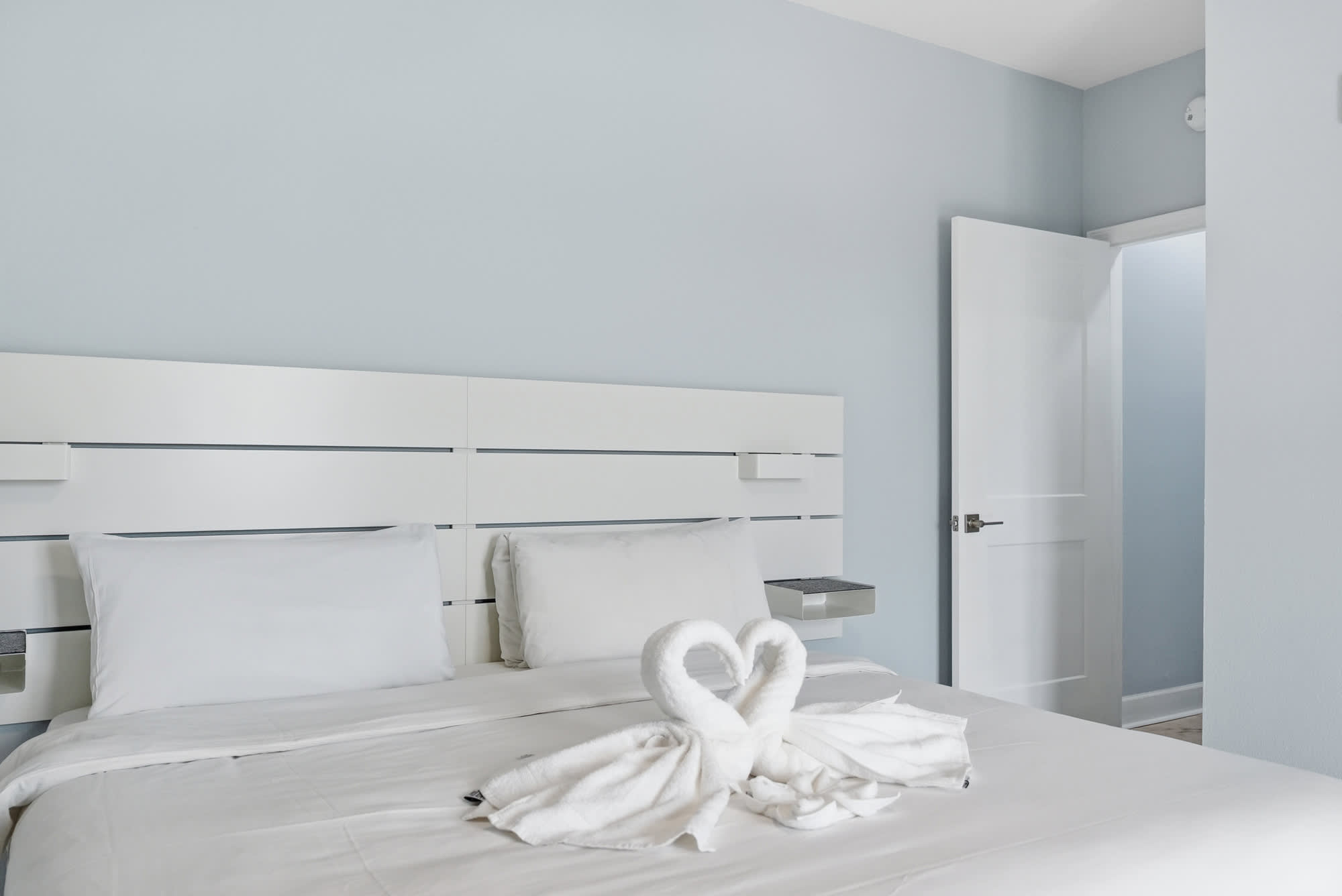 Tidewater 200 – Modern bedroom with a white bed, decorative towel swans, and a light blue wall.