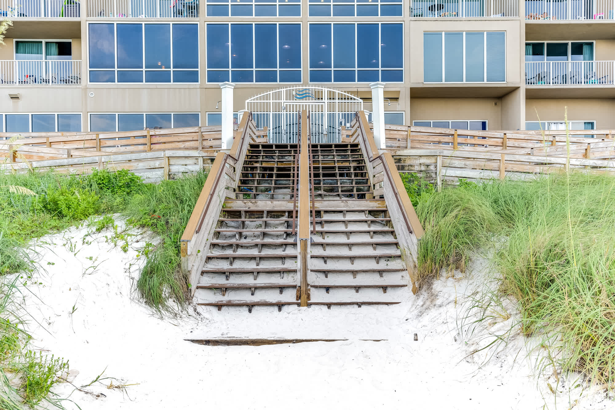 Tidewater 200 – Wooden stairs leading from the beach to a multi-story building with large windows.