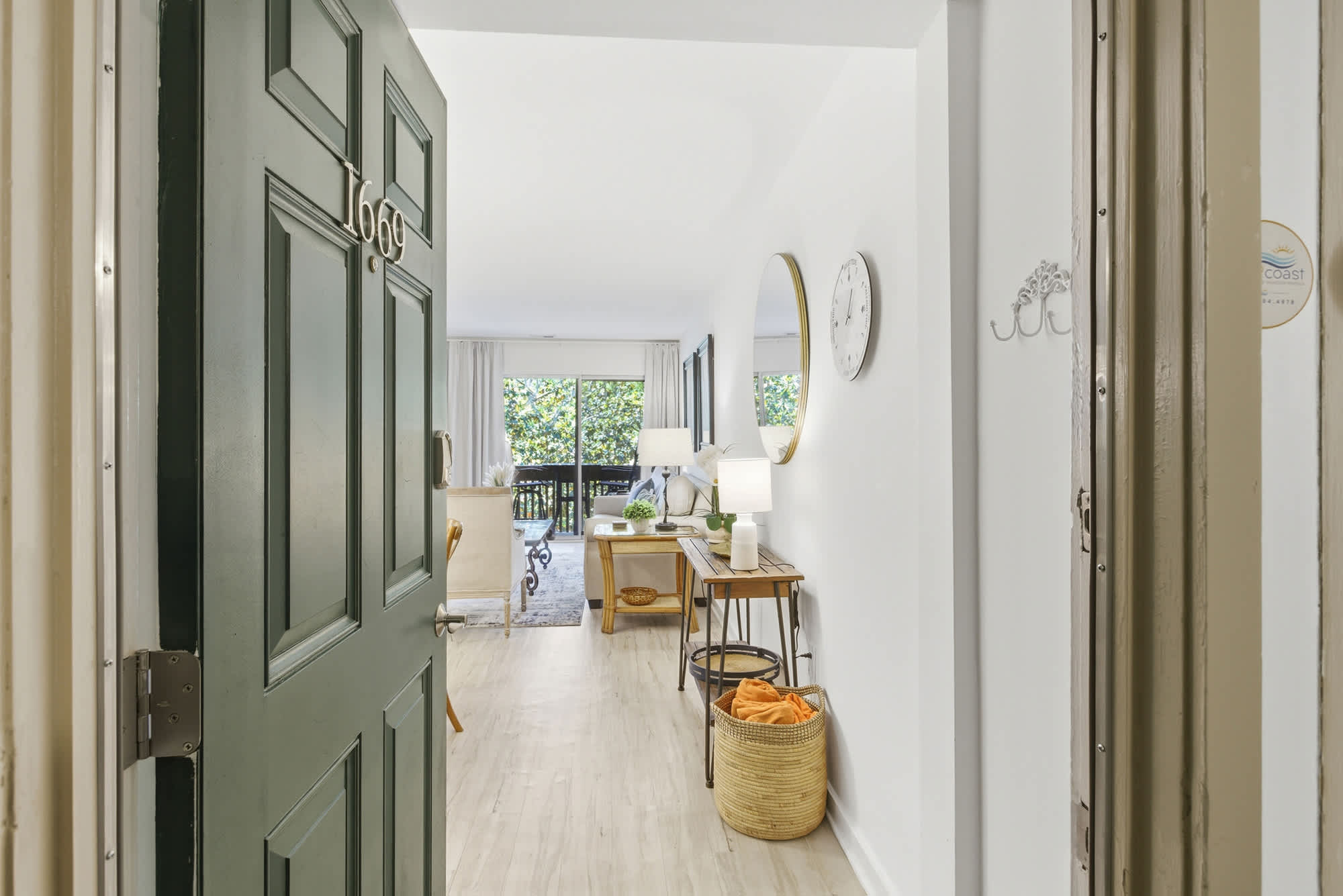 Crisp white walls and honey-toned floors guide your gaze toward a bright living area framed by lush greenery. Woven textures and soft lamp glows create an immediate sense of calm as you cross the threshold into this airy, modern home.