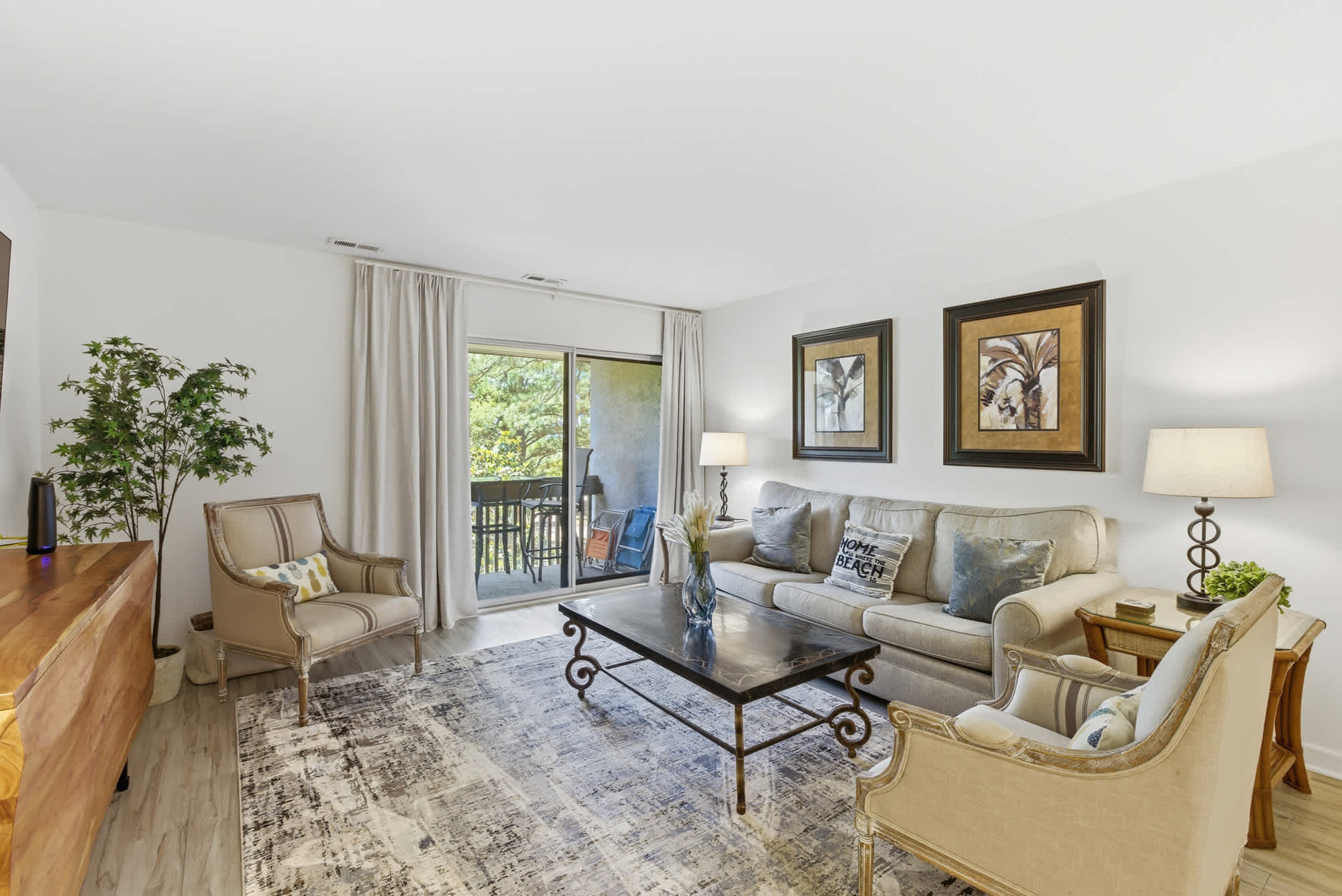 Creamy linen upholstery and vintage-inspired chairs create a soft, communal space for quiet afternoons. Natural light spills through sliding glass doors, highlighting the rich wood grain and textured rug that ground this airy coastal living room.