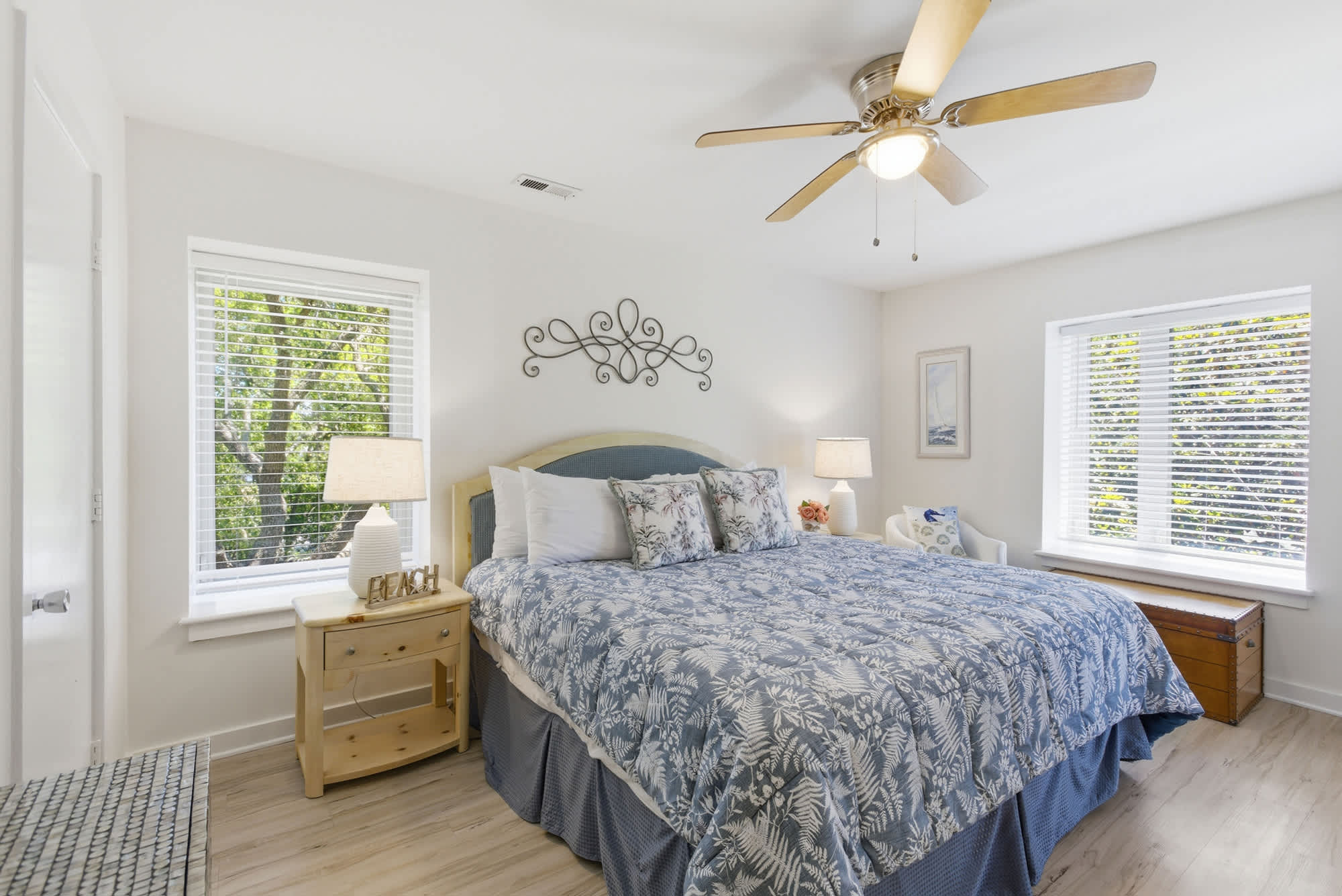 Floral patterns and soft blue linens create a serene escape in this airy bedroom. Natural light filters through the blinds to illuminate the warm pine furniture and smooth wood flooring for a peaceful morning.