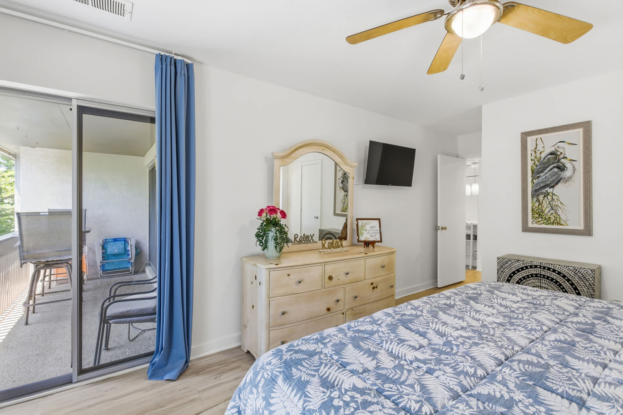 Azure blue curtains frame a large sliding door, inviting the afternoon glow into this airy bedroom. Natural pine furniture and botanical prints create a serene, coastal atmosphere perfect for a restful morning.