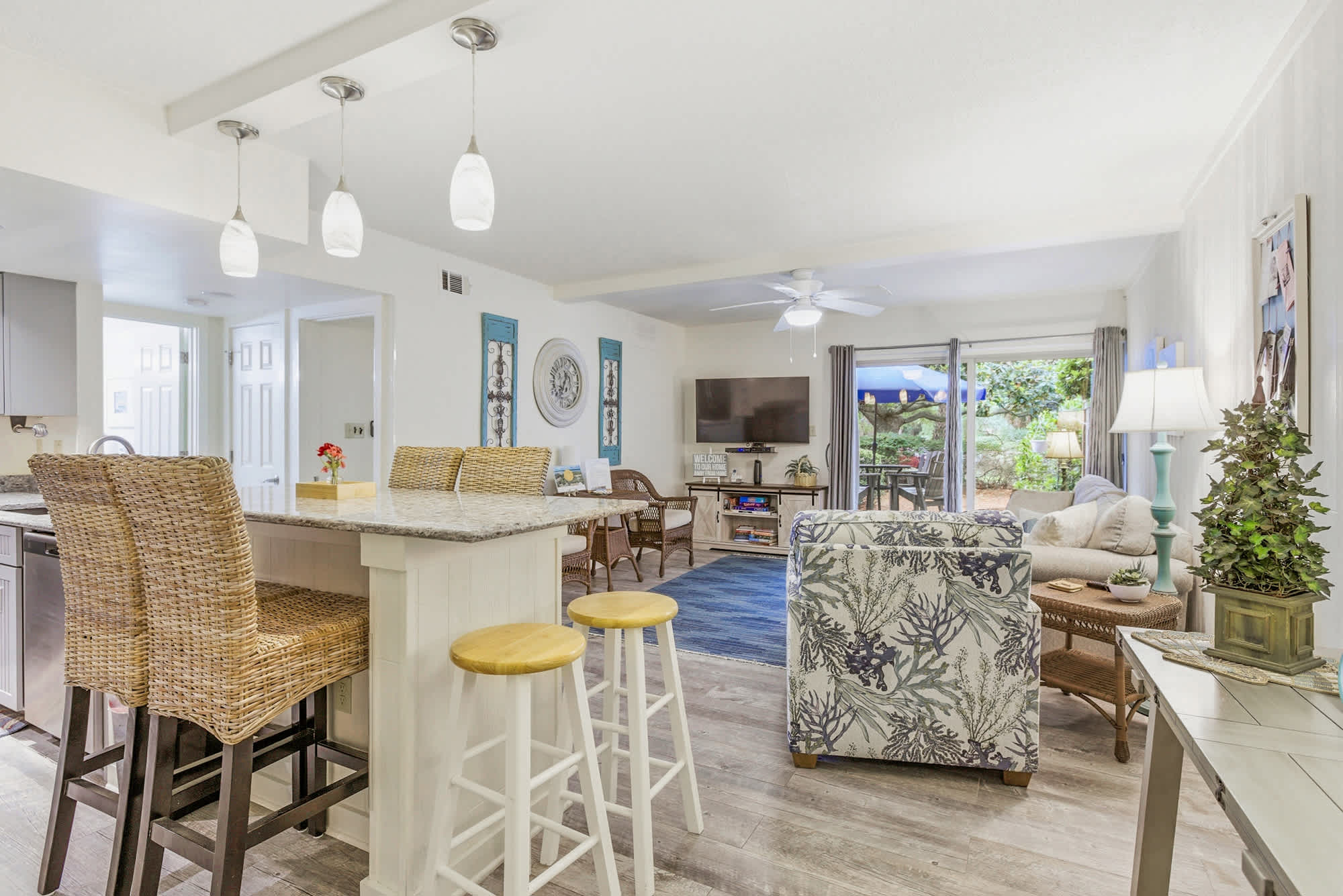 Woven textures and sea-inspired prints create a breezy, coastal atmosphere in this open living area. Natural light pours through the glass doors, illuminating the granite island and inviting you to gather for coffee before a day at the beach.